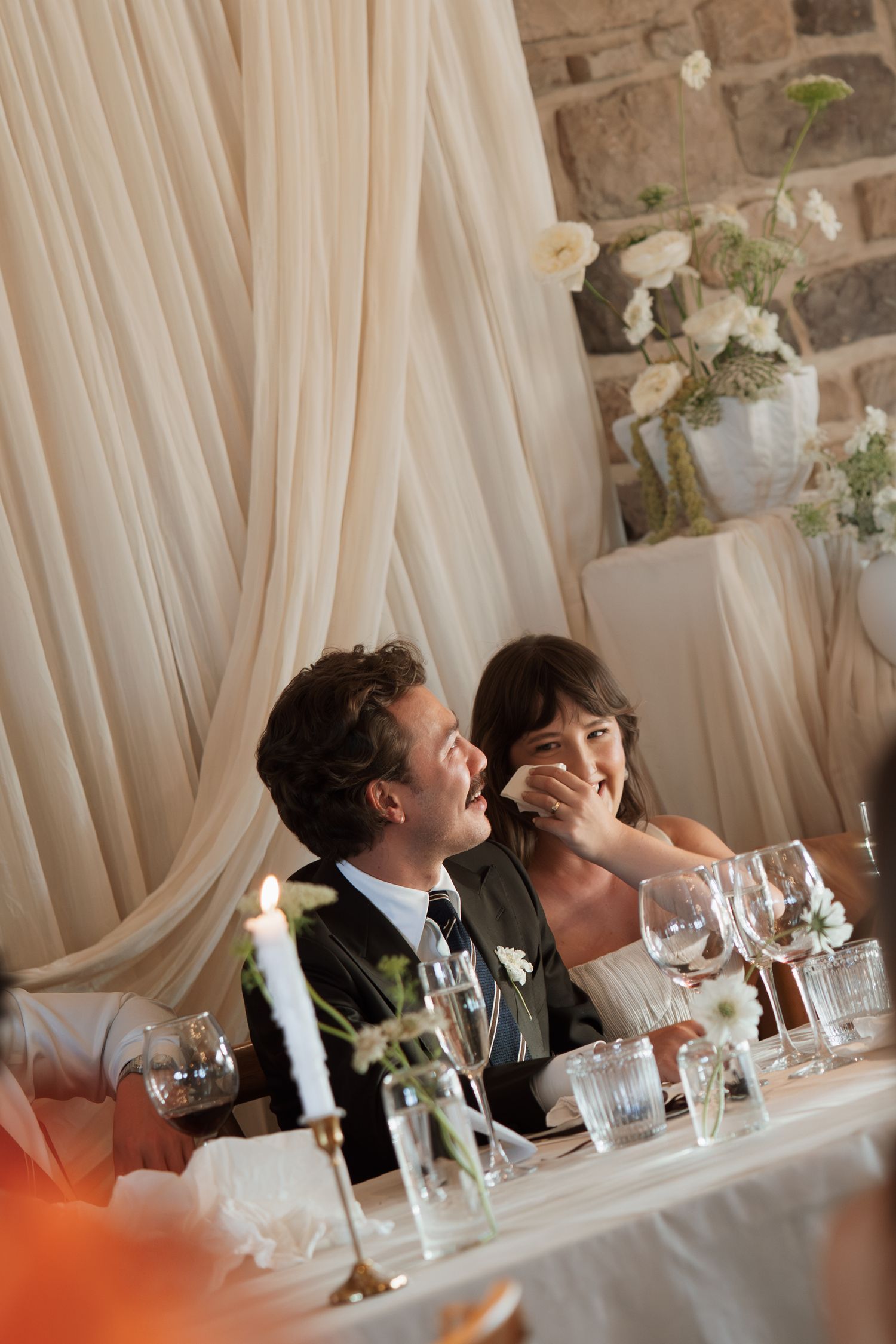 Candid moments of celebration at wedding reception head table with white draping.