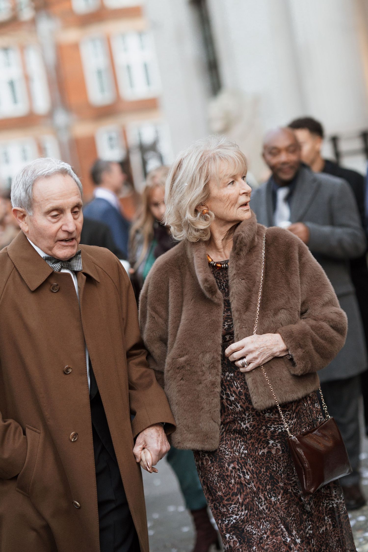 An elderly couple in winter coats walking together at an event.