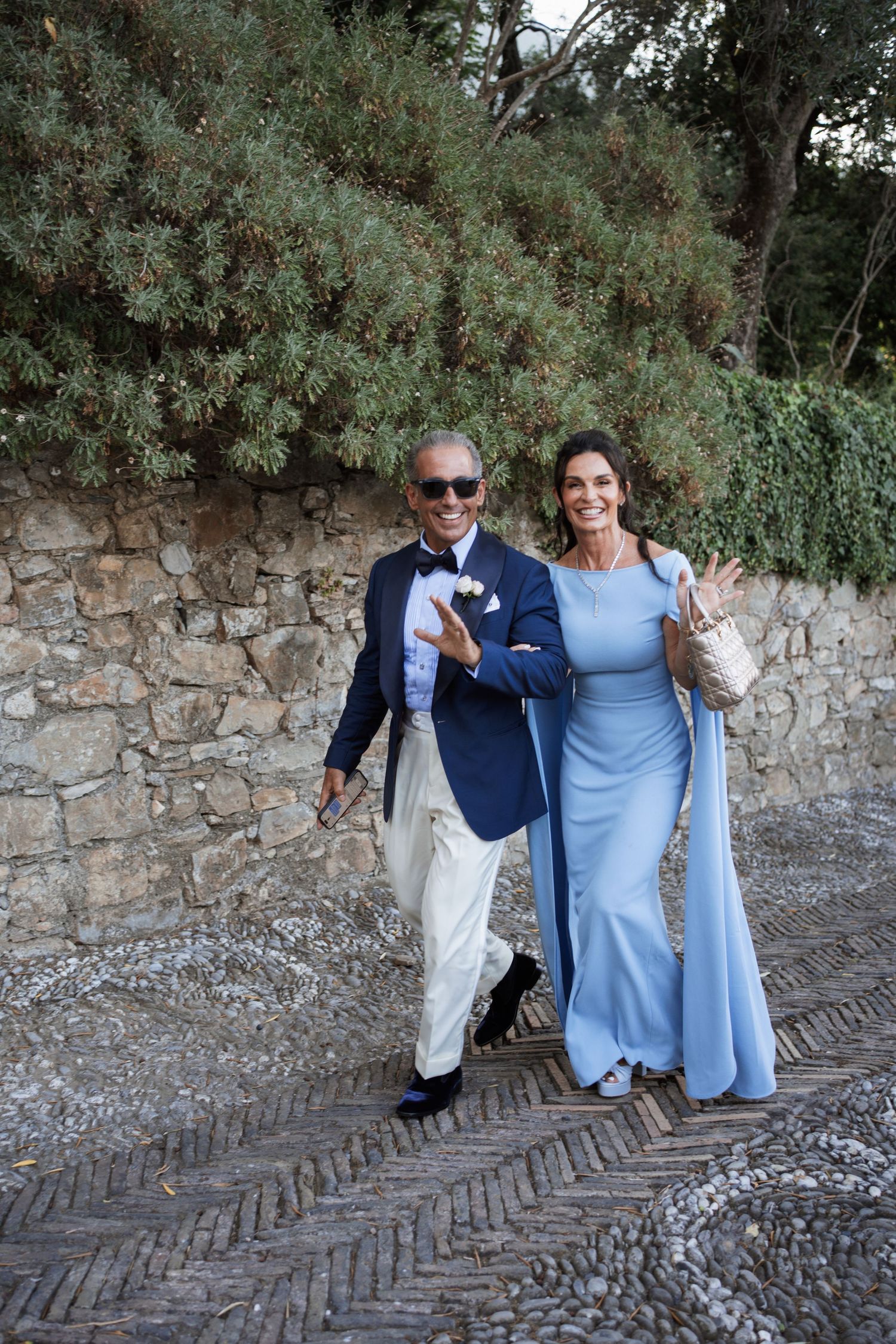 Elegantly dressed couple walking along a stone path at a formal outdoor event.
