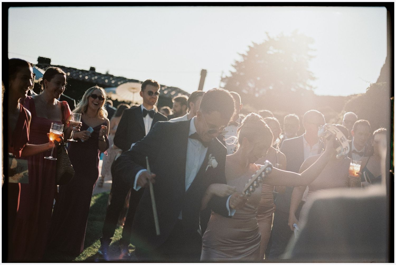Guests in formal attire celebrate outdoors at a golden hour wedding reception with warm sunlight glowing in background.