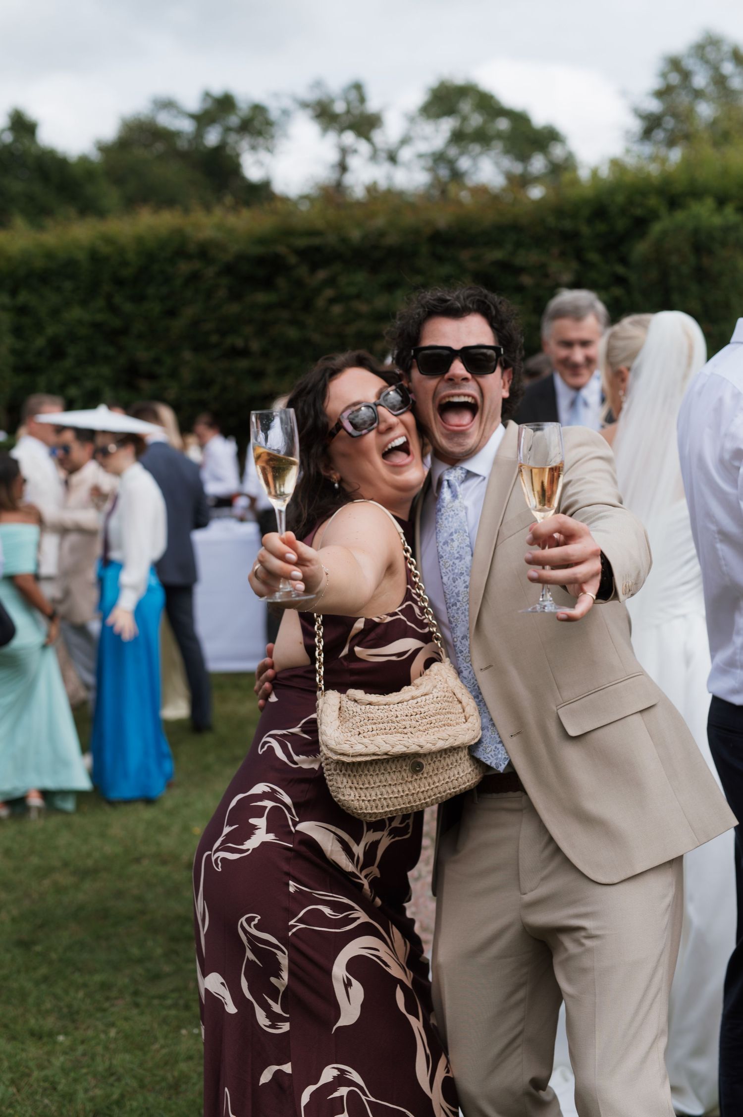 Two laughing wedding guests in sunglasses toast with champagne glasses outdoors at a garden wedding reception.