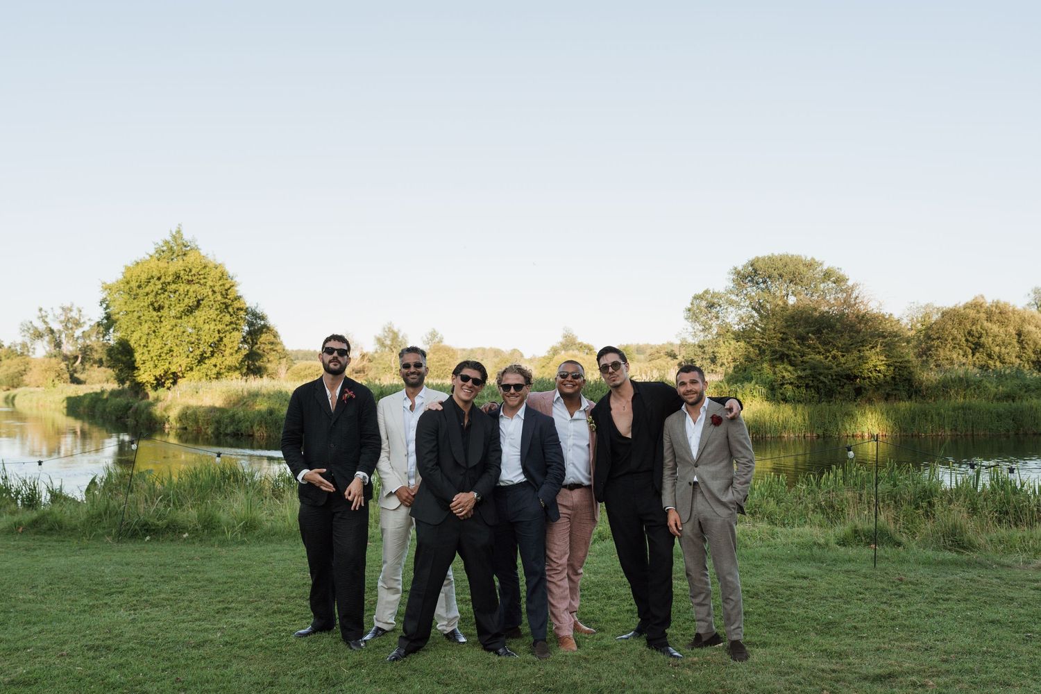 Group of eight men posing outdoors near a pond, dressed in formal attire, surrounded by lush green trees.