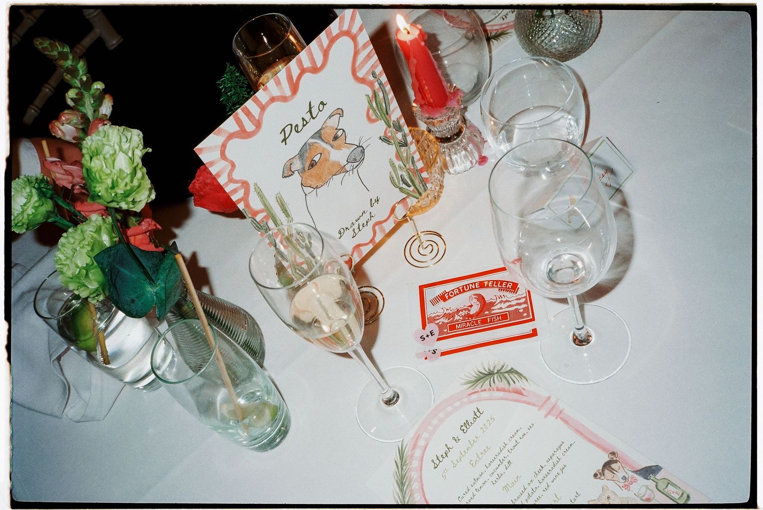 Elegant wedding table setting with floral arrangements, wine glasses, candles, and decorative place cards on white tablecloth.