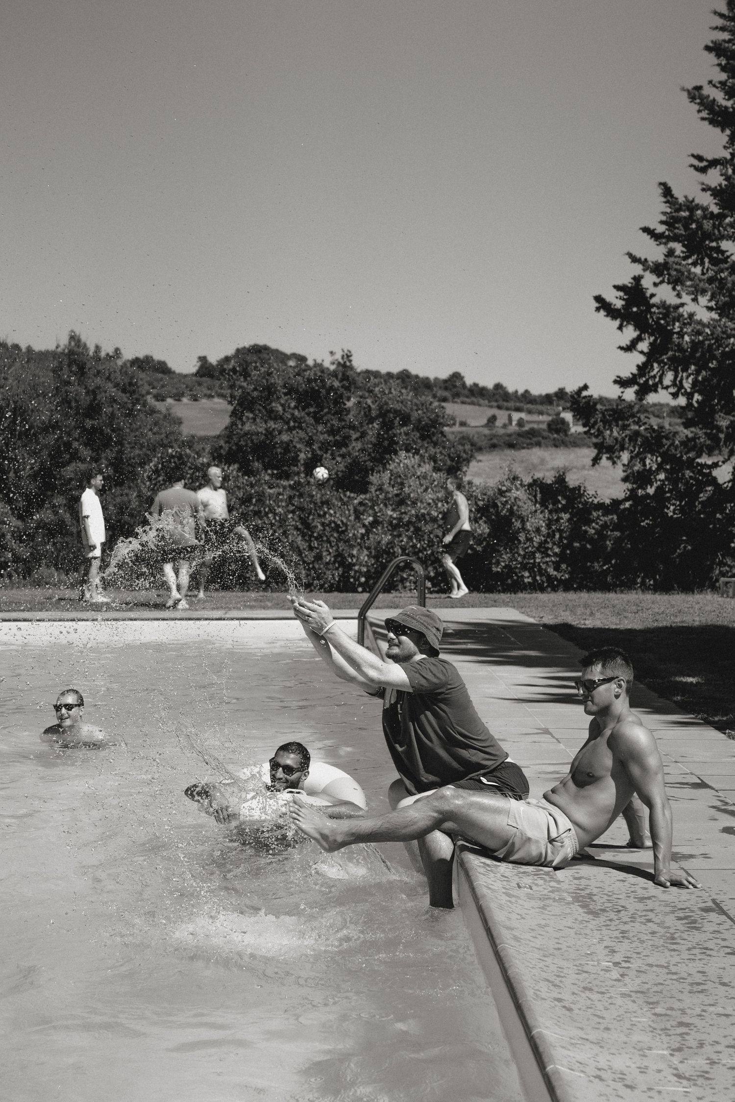 Black and white photo of men playing and swimming in an outdoor pool surrounded by lush trees on a sunny day.