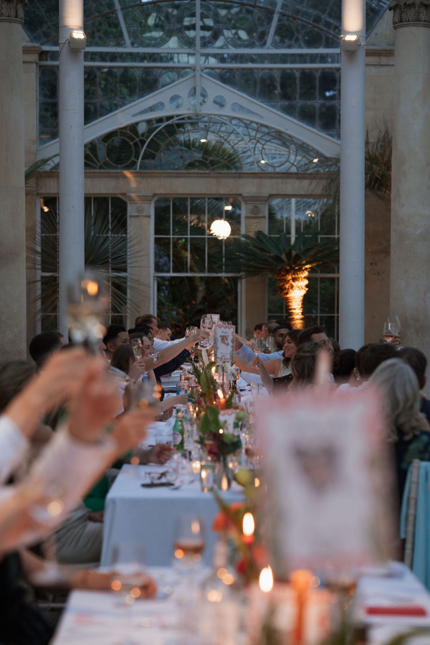 Evening wedding reception with guests toasting under a glass ceiling with ambient lighting.