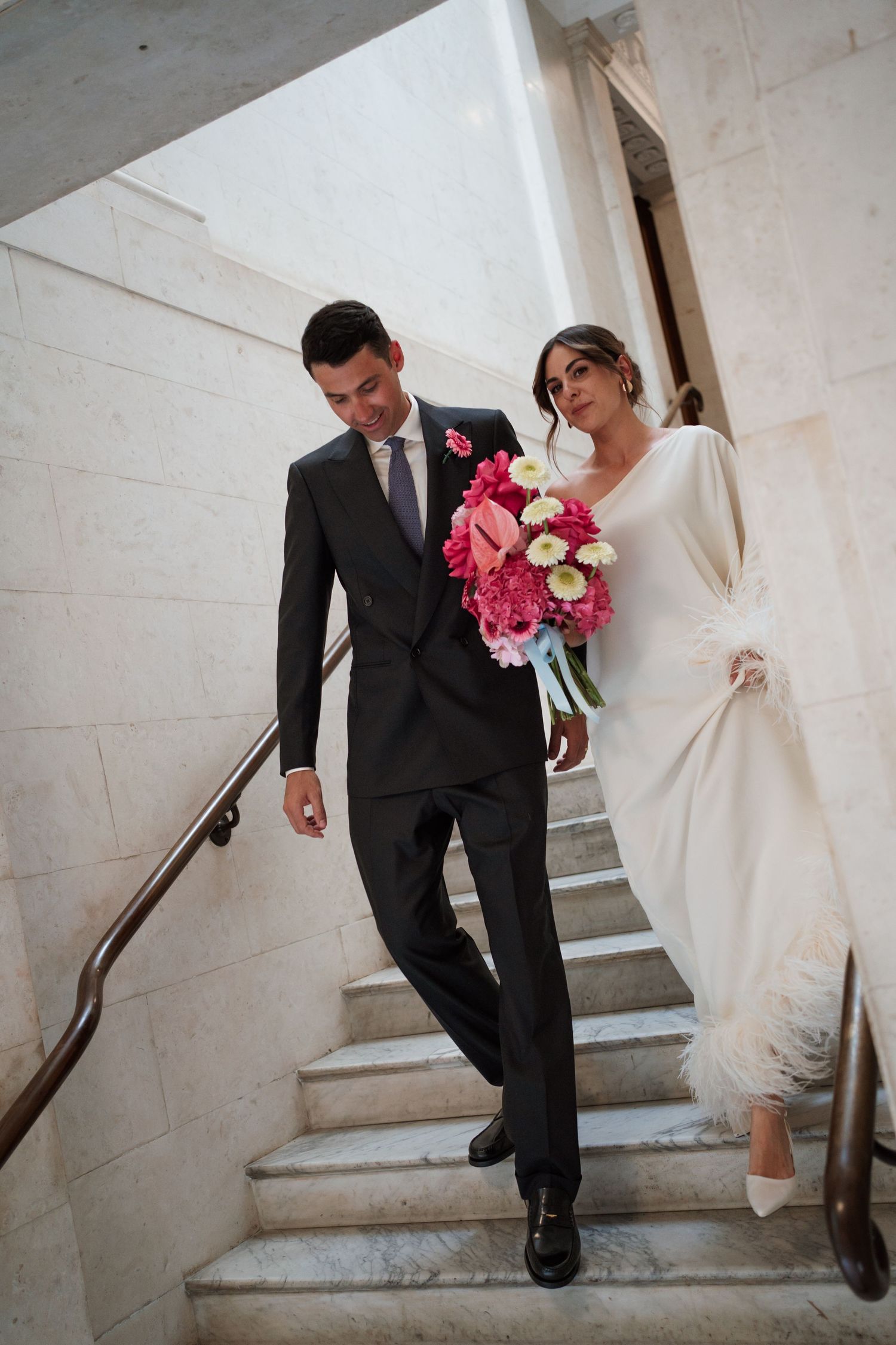 Wedding couple descends white marble staircase while holding pink floral bouquet.