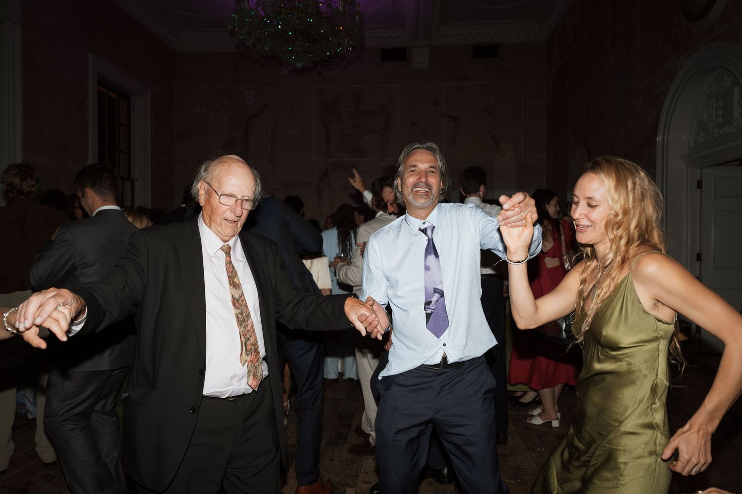 Three guests hold hands and dance joyfully at an indoor evening party, two men in dress shirts and ties and a woman in an olive satin dress.