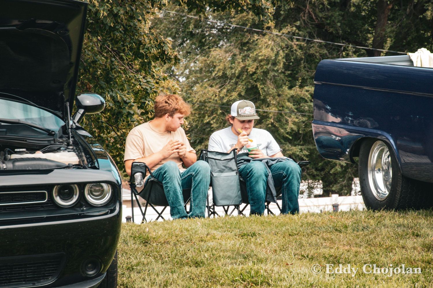 Two people sitting in camping chairs at a car show or outdoor event.