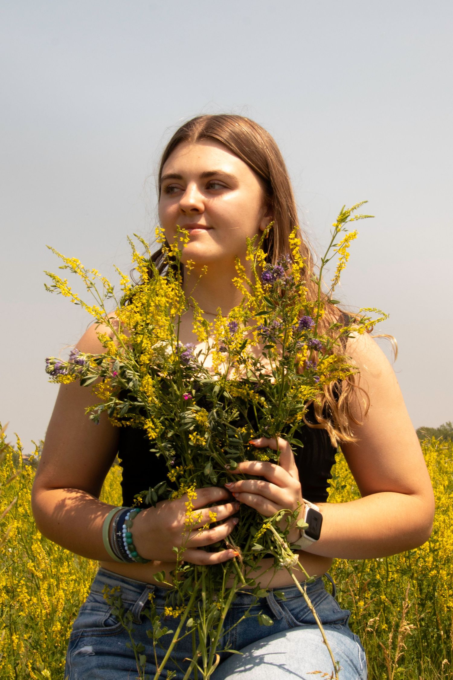 A person holding a bouquet of yellow wildflowers poses in a sunny field.