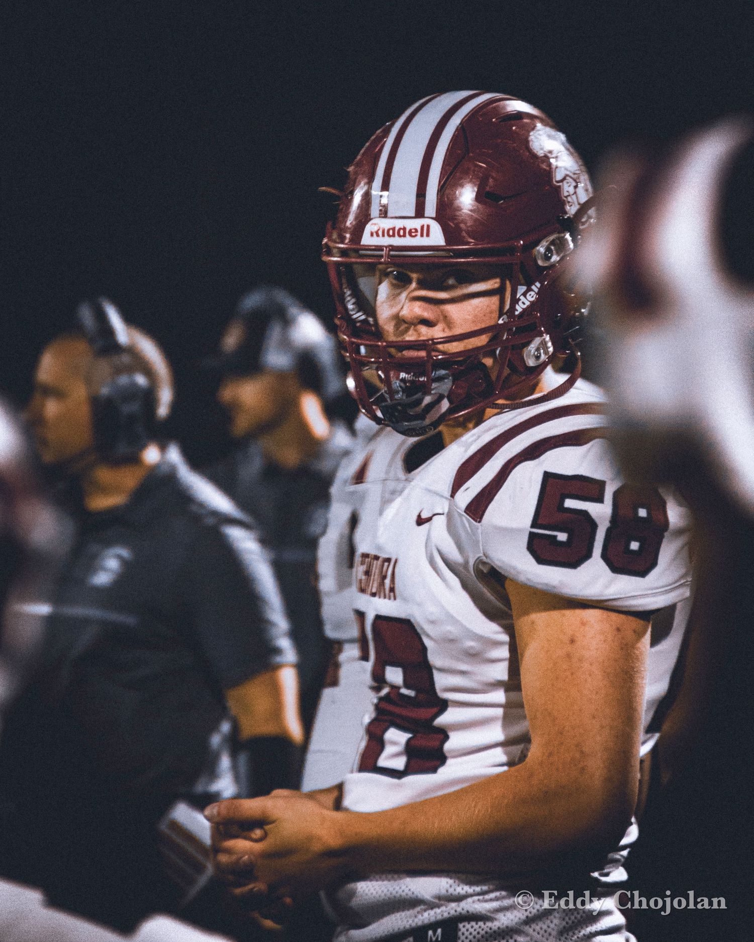 Football players in maroon and white uniforms lined up on sideline during nighttime game.