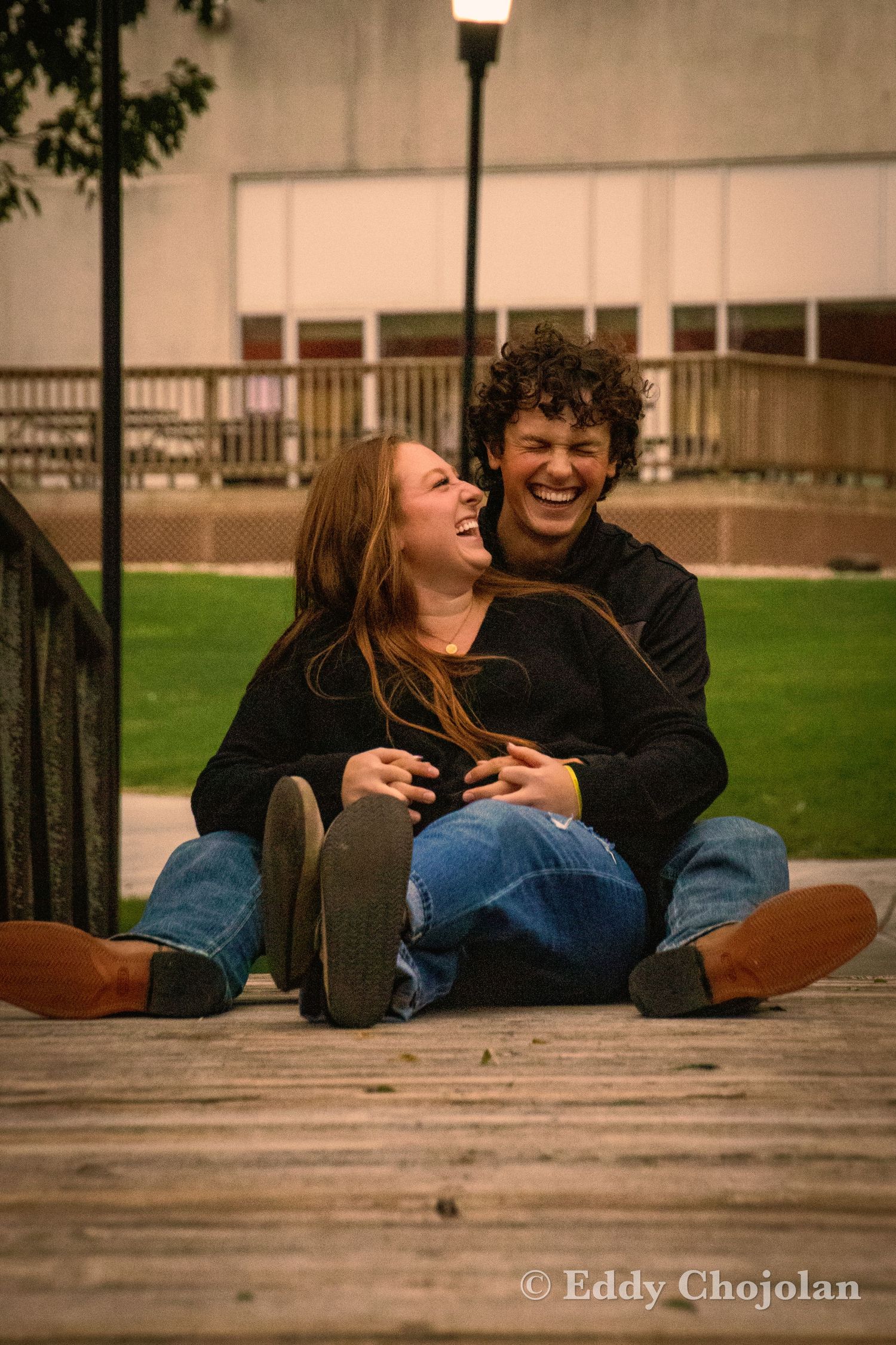 A playful couple laughs together while relaxing on wooden decking outdoors.