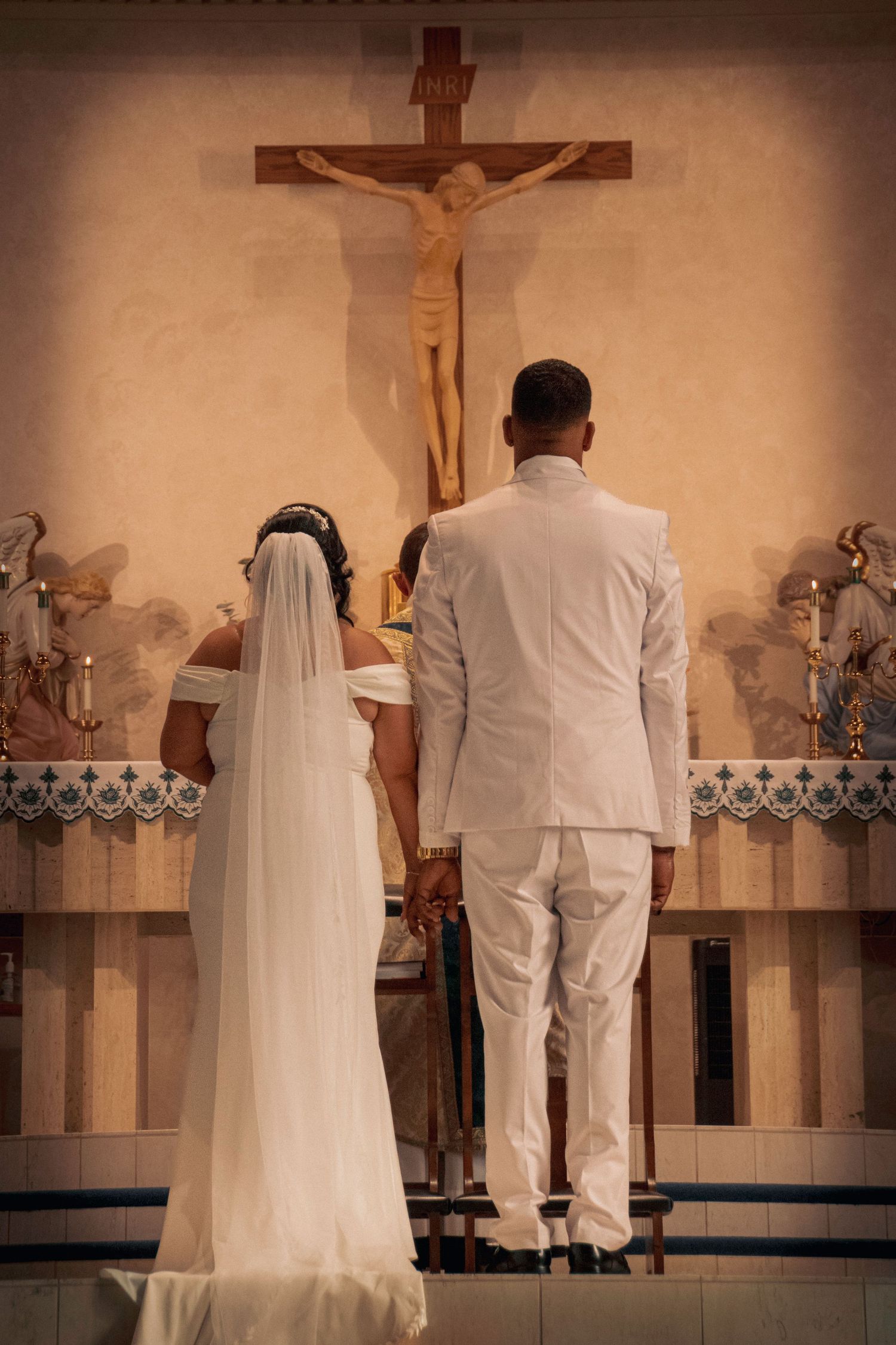 A couple in white attire stands before a church altar with a crucifix during a wedding ceremony.