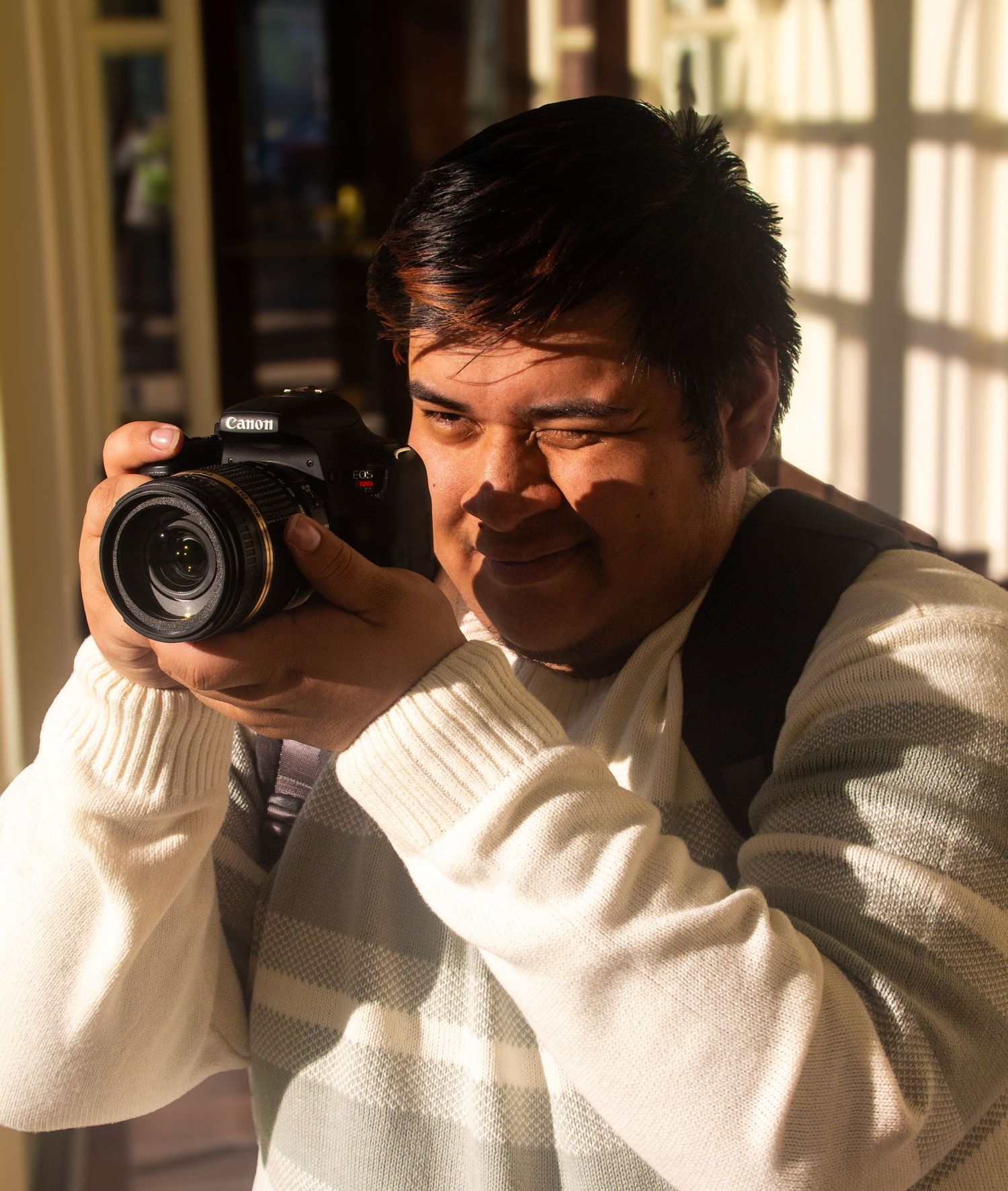 A photographer in a white sweater and dark vest holds a professional camera while taking a photo in natural lighting.