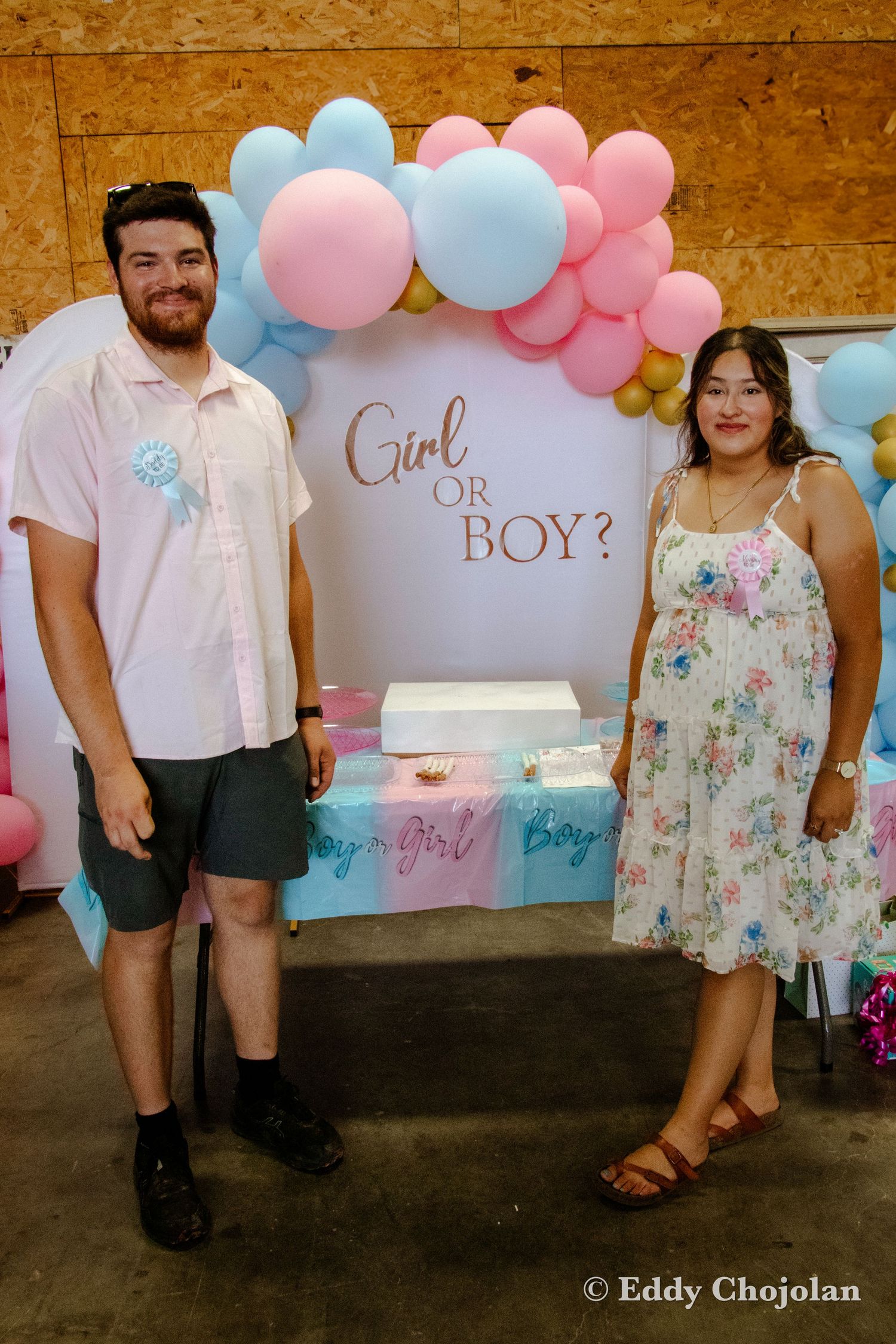 Expectant couple standing by 'Girl or Boy' sign with pink and blue balloon decorations.