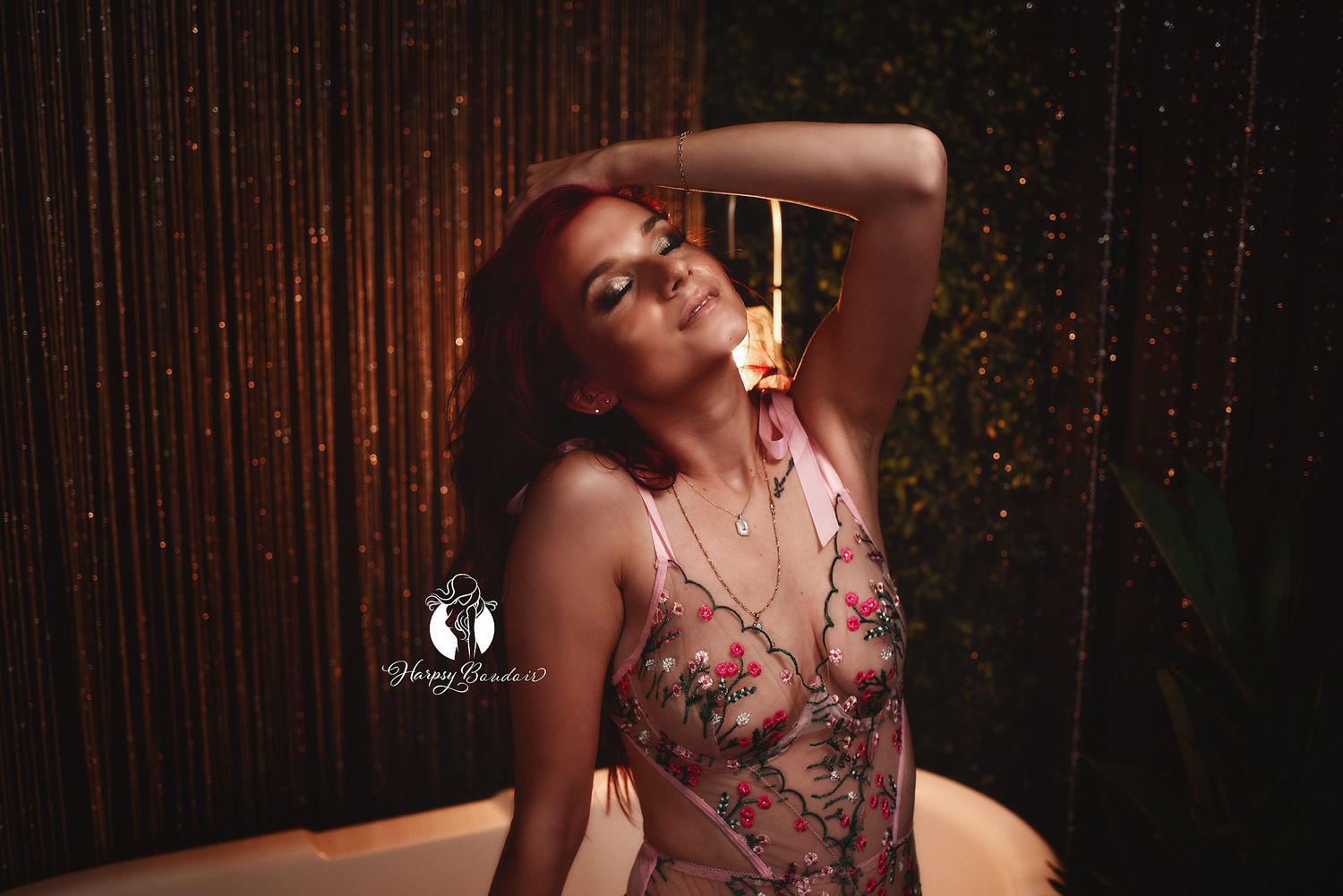 Sexy boudoir client with red hair, wearing sheer pink lingerie with flowers poses with hand in hair and back arched in boudoir photoshoot