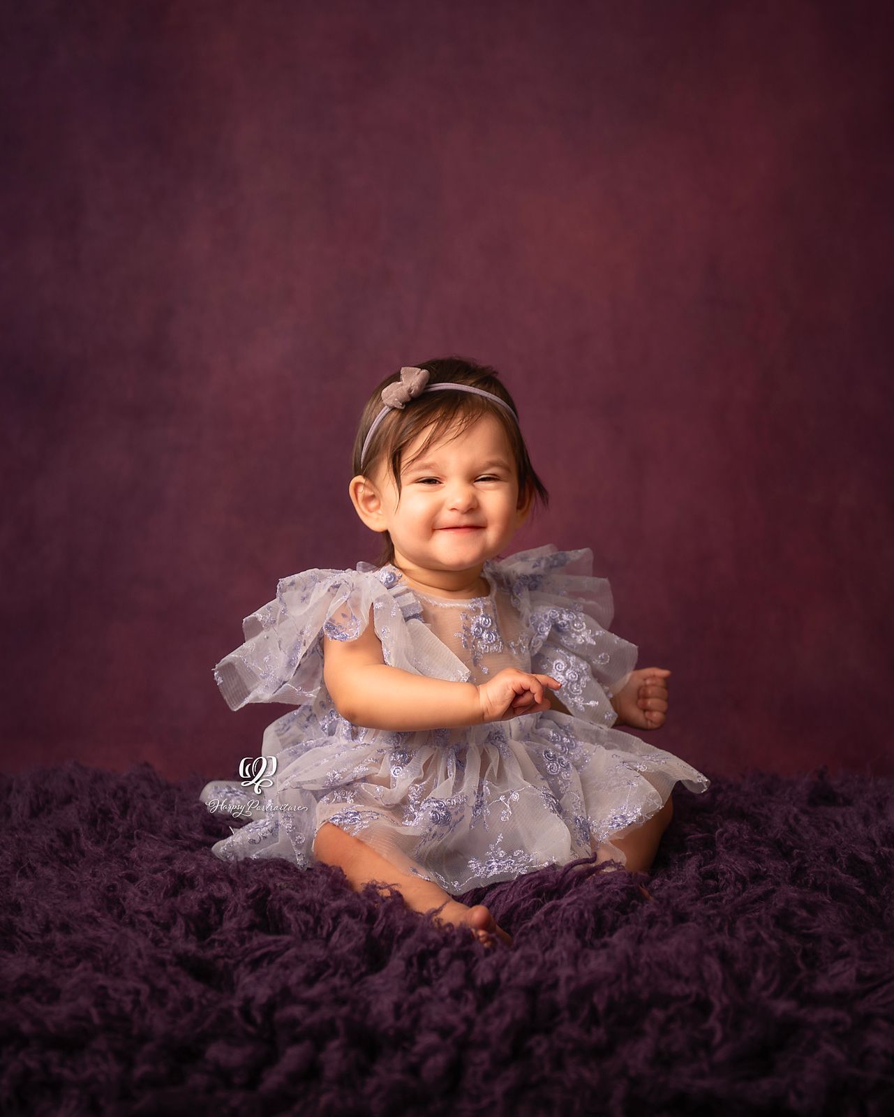 Baby girl wearing purple poofy dress with purple backdrop& fur rug smiling at camera during Sitter Session at Lexington,
