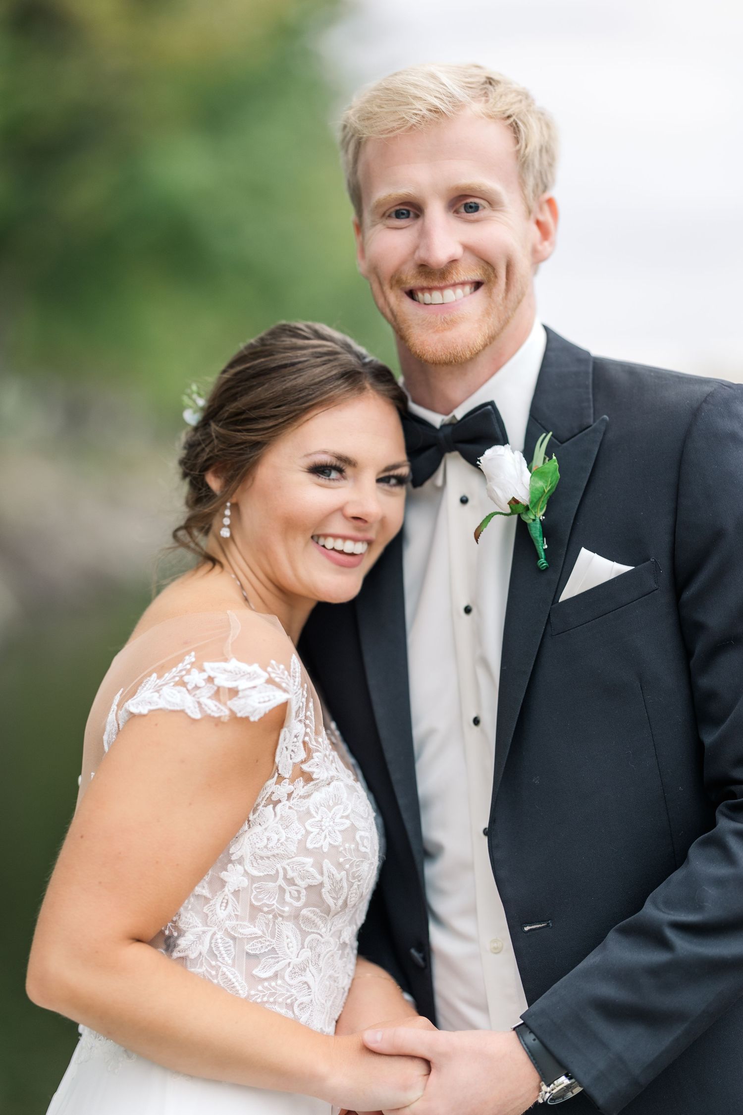 A bride in lace dress and groom in tuxedo share a loving embrace outdoors.