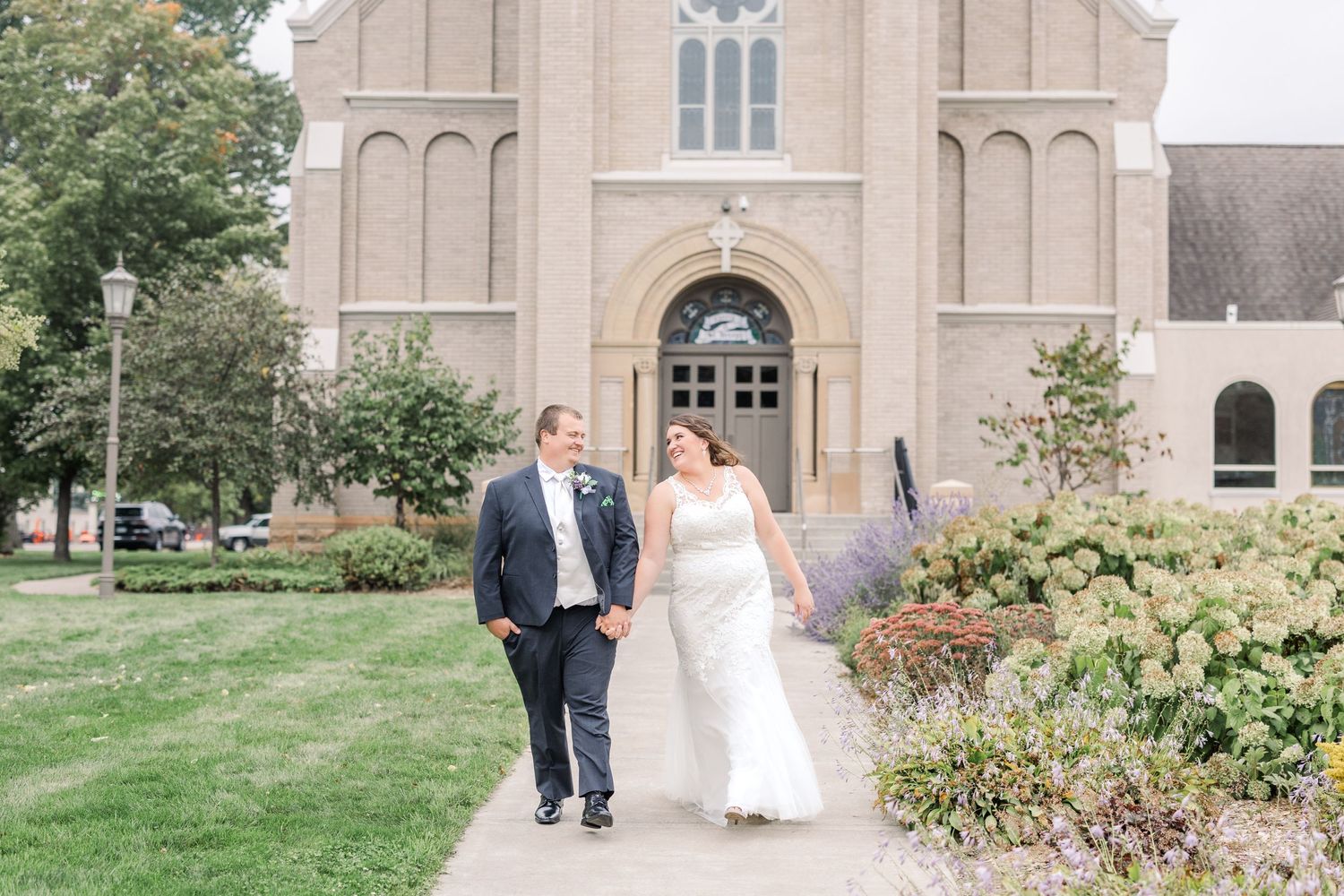 A couple exits a beautiful stone church surrounded by manicured gardens and greenery.