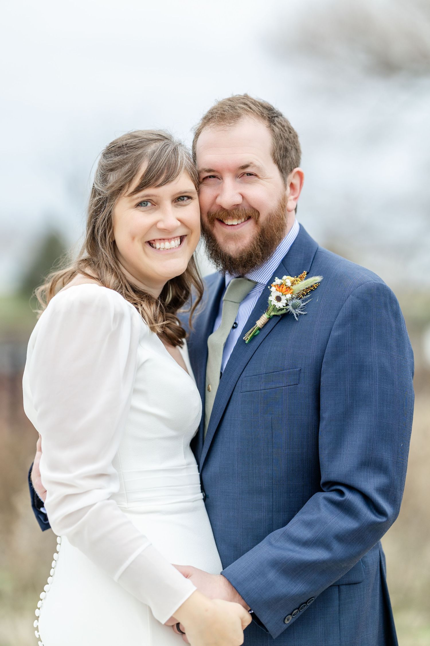 A joyful wedding couple embraces and smiles outdoors while wearing a white dress and navy blue suit.