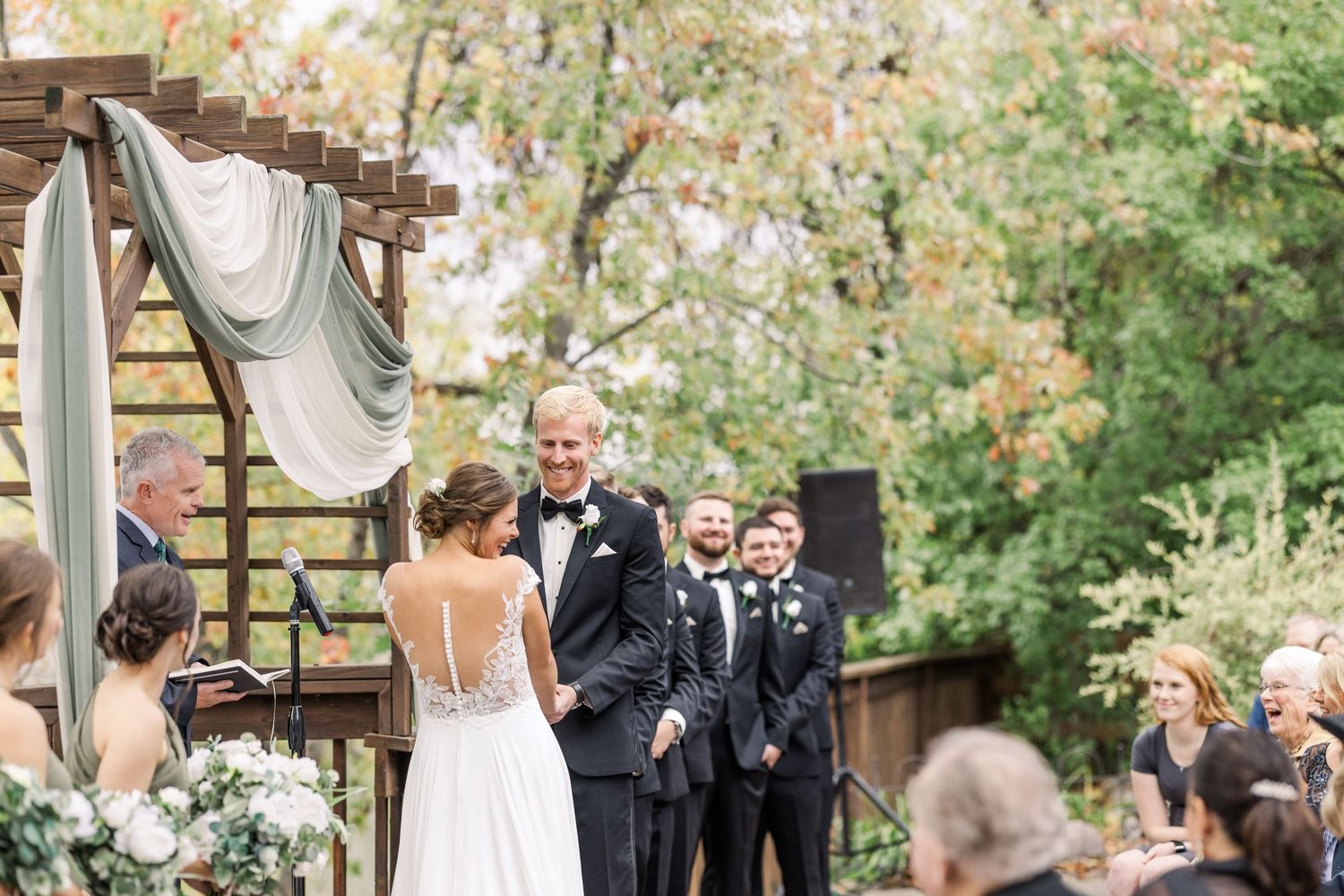 A beautiful outdoor wedding ceremony under a wooden arch decorated with white fabric and flowers.