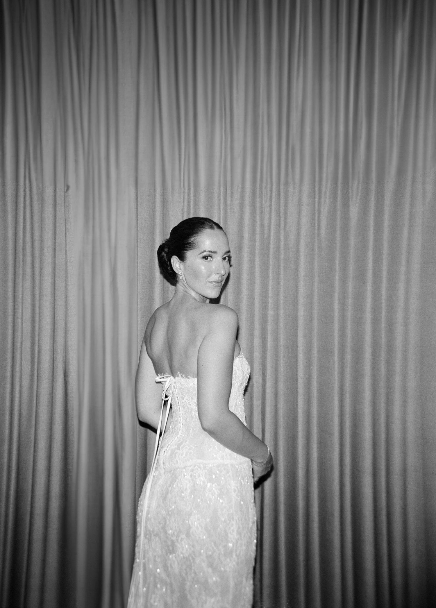 Black and white photo of a woman in a strapless lace gown, looking over her shoulder against draped curtains.