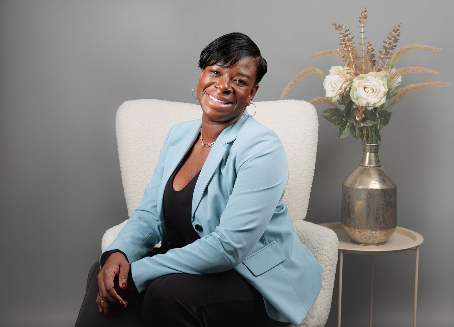 Professional wearing light blue blazer poses confidently against gray backdrop with floral decor.