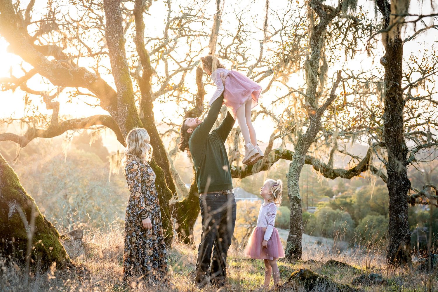 Family enjoying golden hour outdoors, father lifting child in air near oak trees while mother and sibling watch.