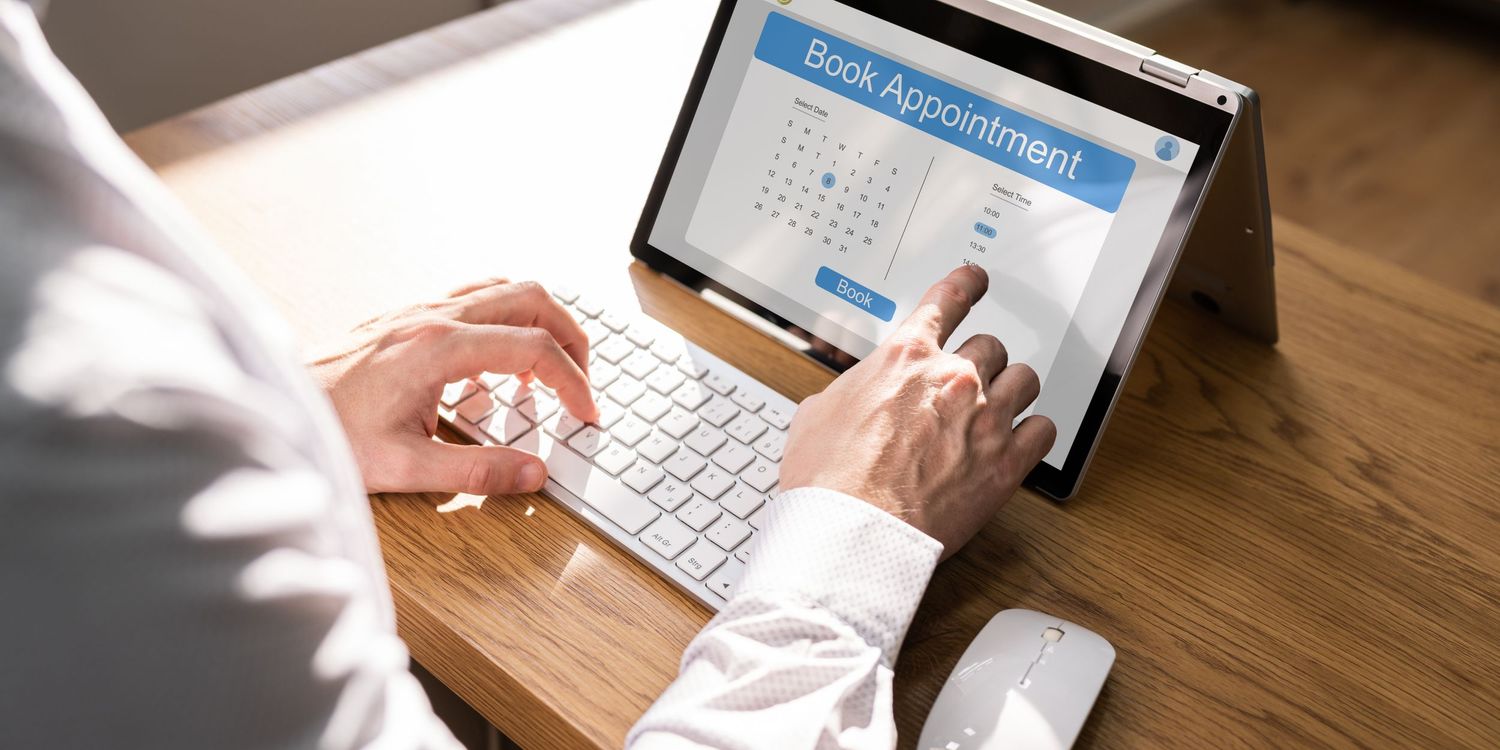 Person in white sleeves typing on laptop keyboard with tablet device display showing calendar interface on wooden desk.