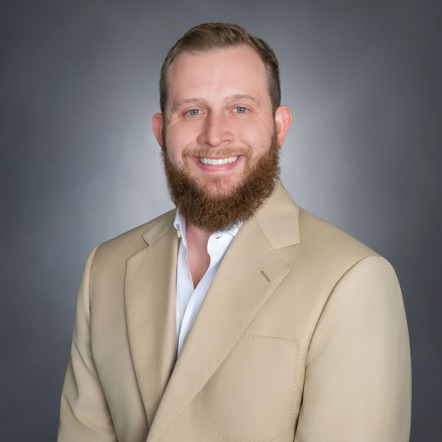 Professional headshot of a smiling man with a beard wearing a tan blazer and white shirt against a gray background.