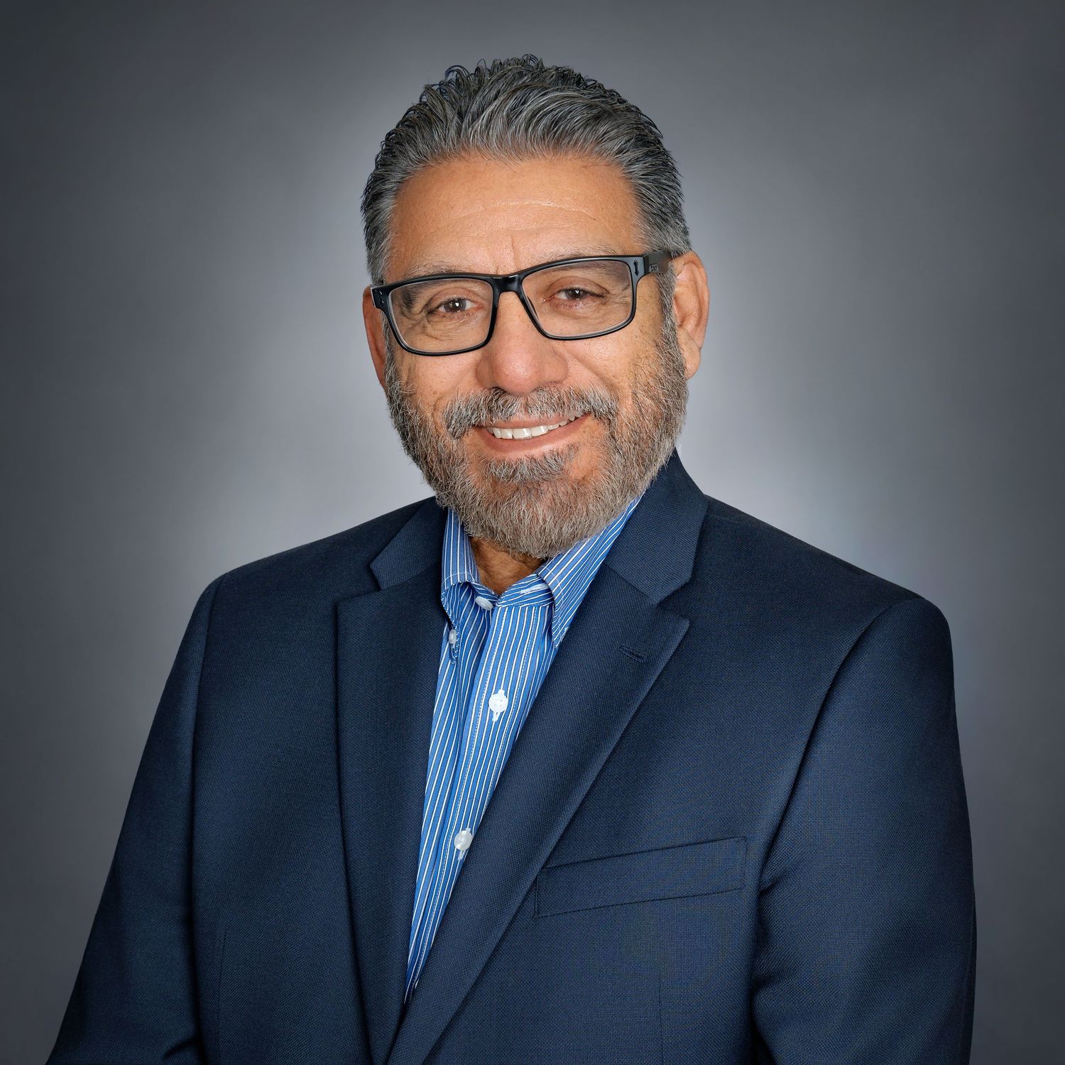Professional headshot of a middle-aged man with glasses and gray hair wearing a navy blazer and blue shirt.