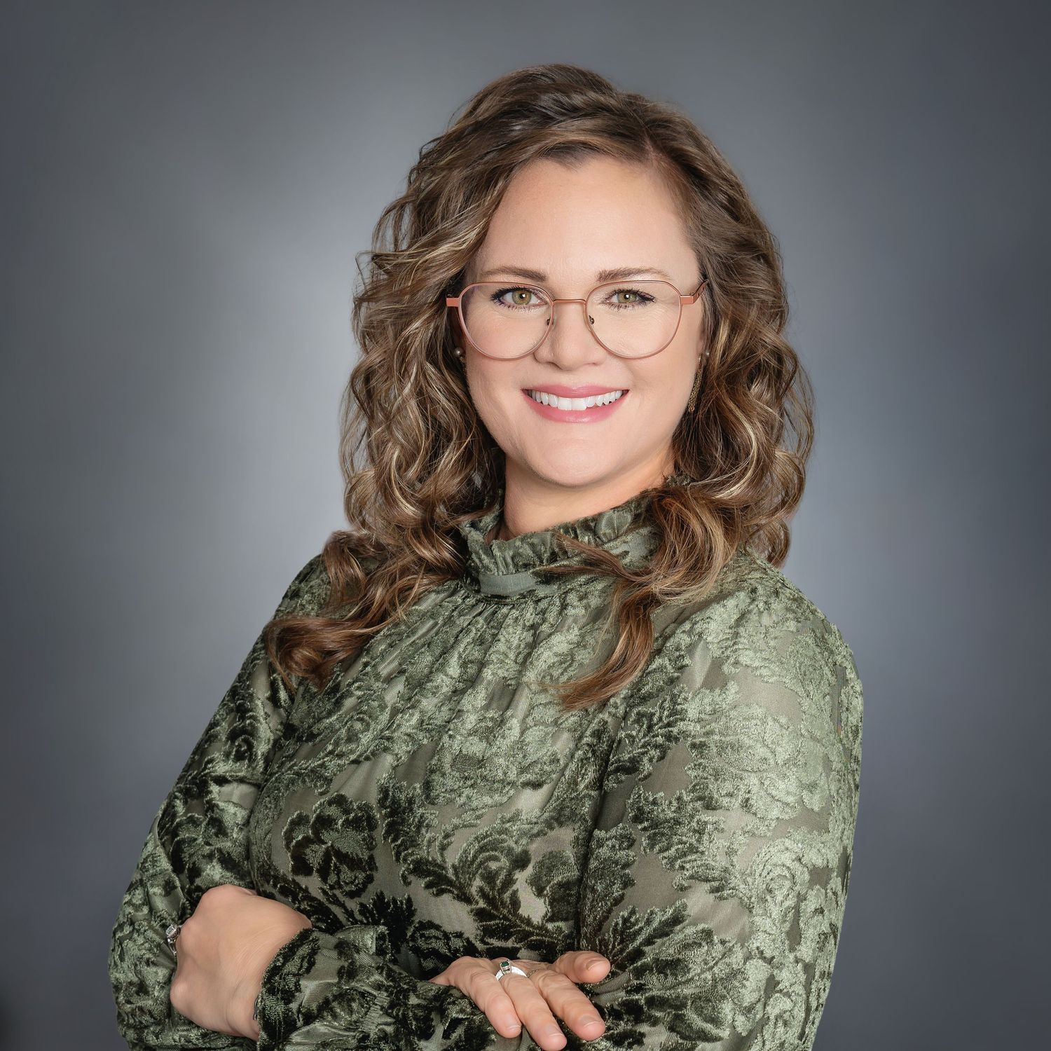 A smiling woman with curly hair and glasses, wearing a green floral blouse, poses with arms crossed against a gray background.