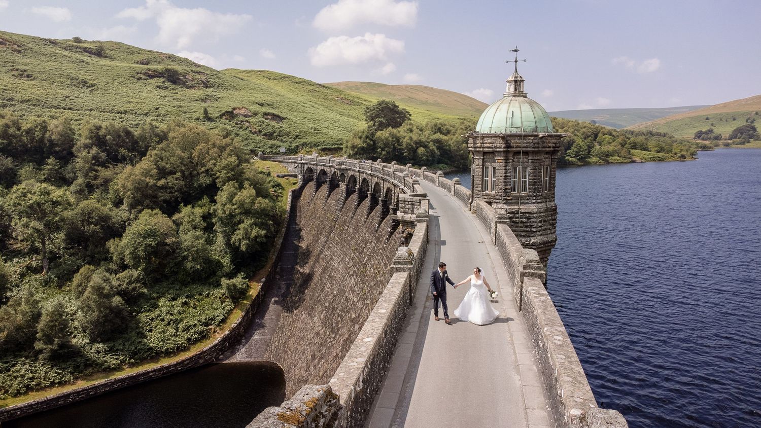 Bride and groom walk along a historic stone dam with a green copper turret, surrounded by lush hills and a serene reservoir.