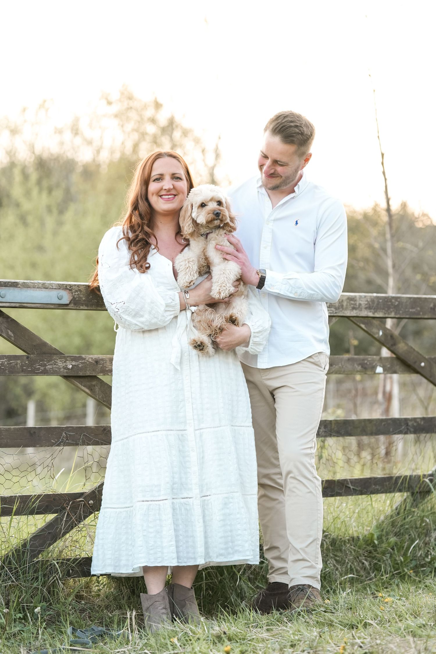 A couple poses outdoors with their small dog near a wooden fence during golden hour.