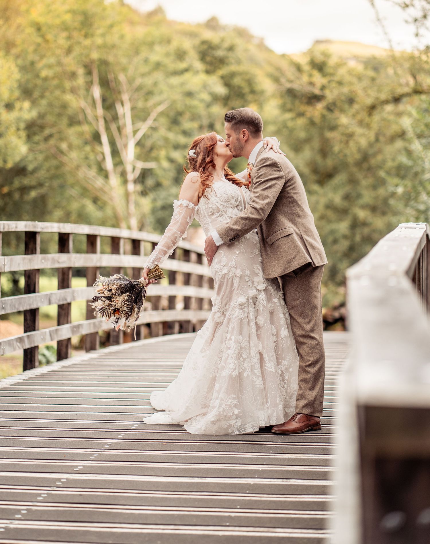 Couple shares romantic kiss on wooden bridge surrounded by lush green trees during outdoor wedding photo session.