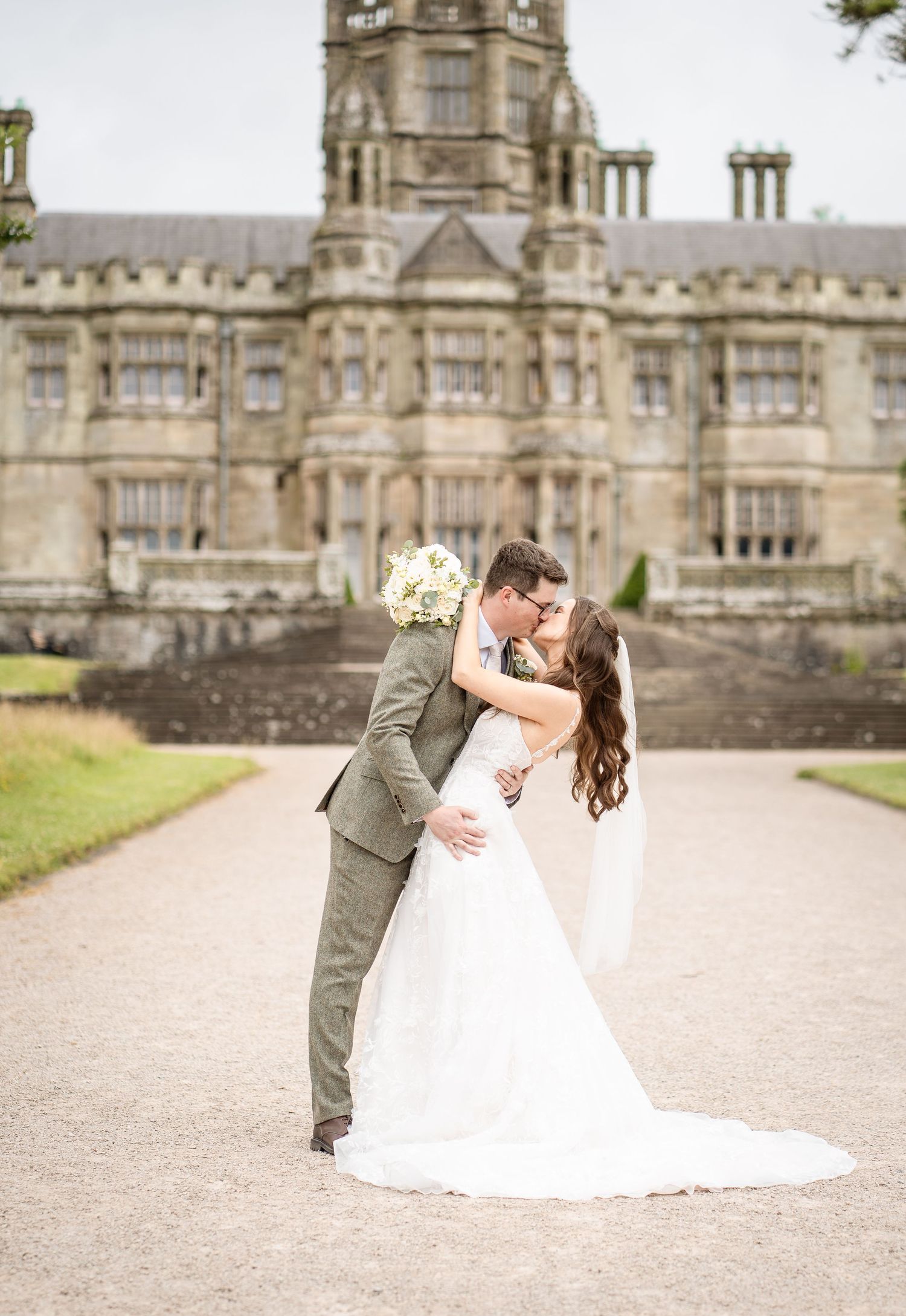 Bride and groom share a romantic dip kiss in front of a grand stone castle on their wedding day.