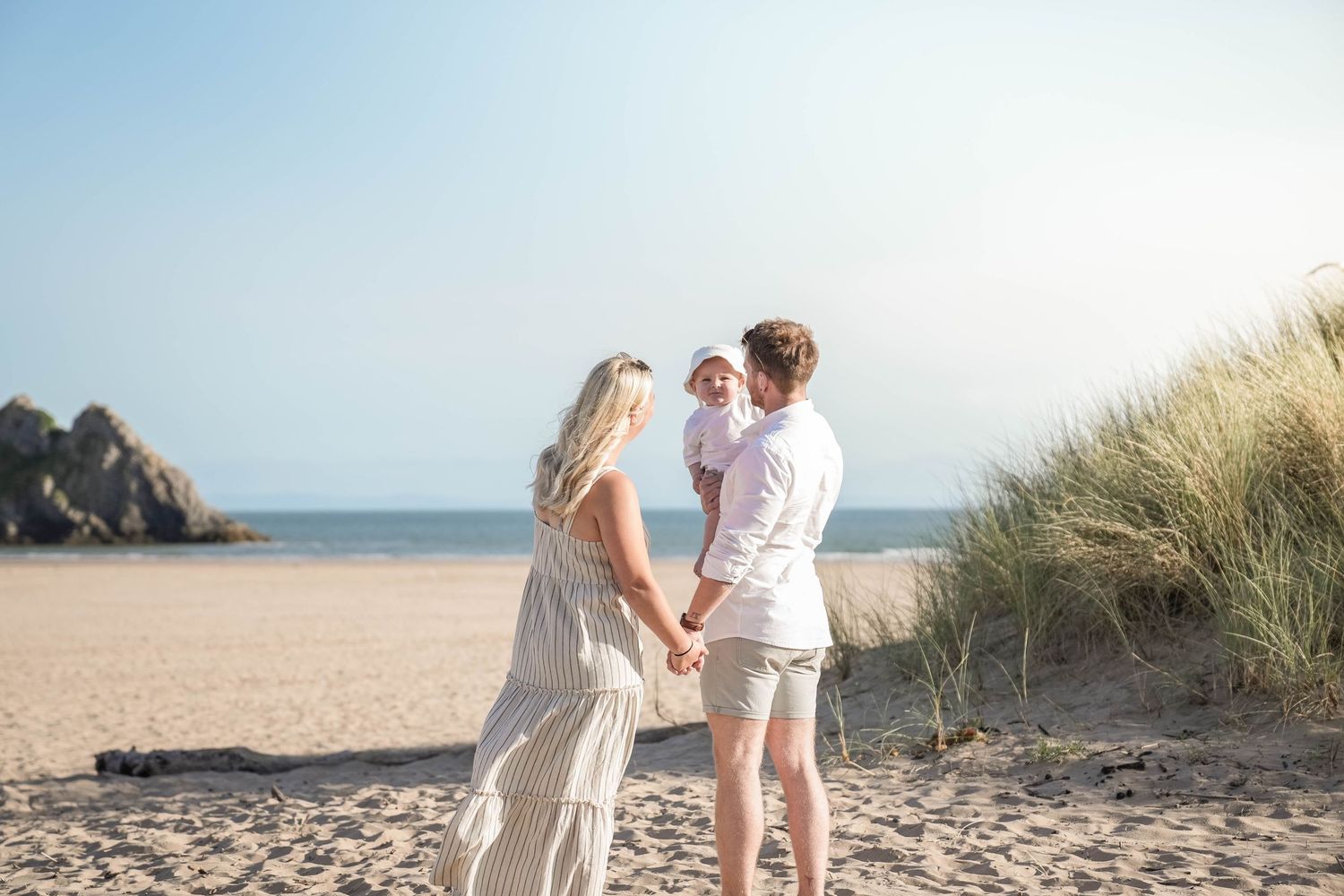 A couple shares a romantic moment together on a sandy beach with tall grass and ocean cliffs in the background.