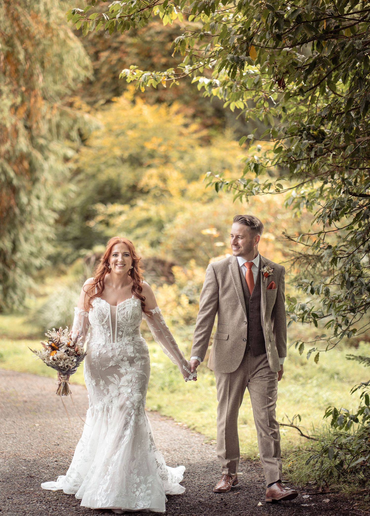 A couple in wedding attire walk hand in hand along a tree-lined path on an autumn day with golden foliage in background.