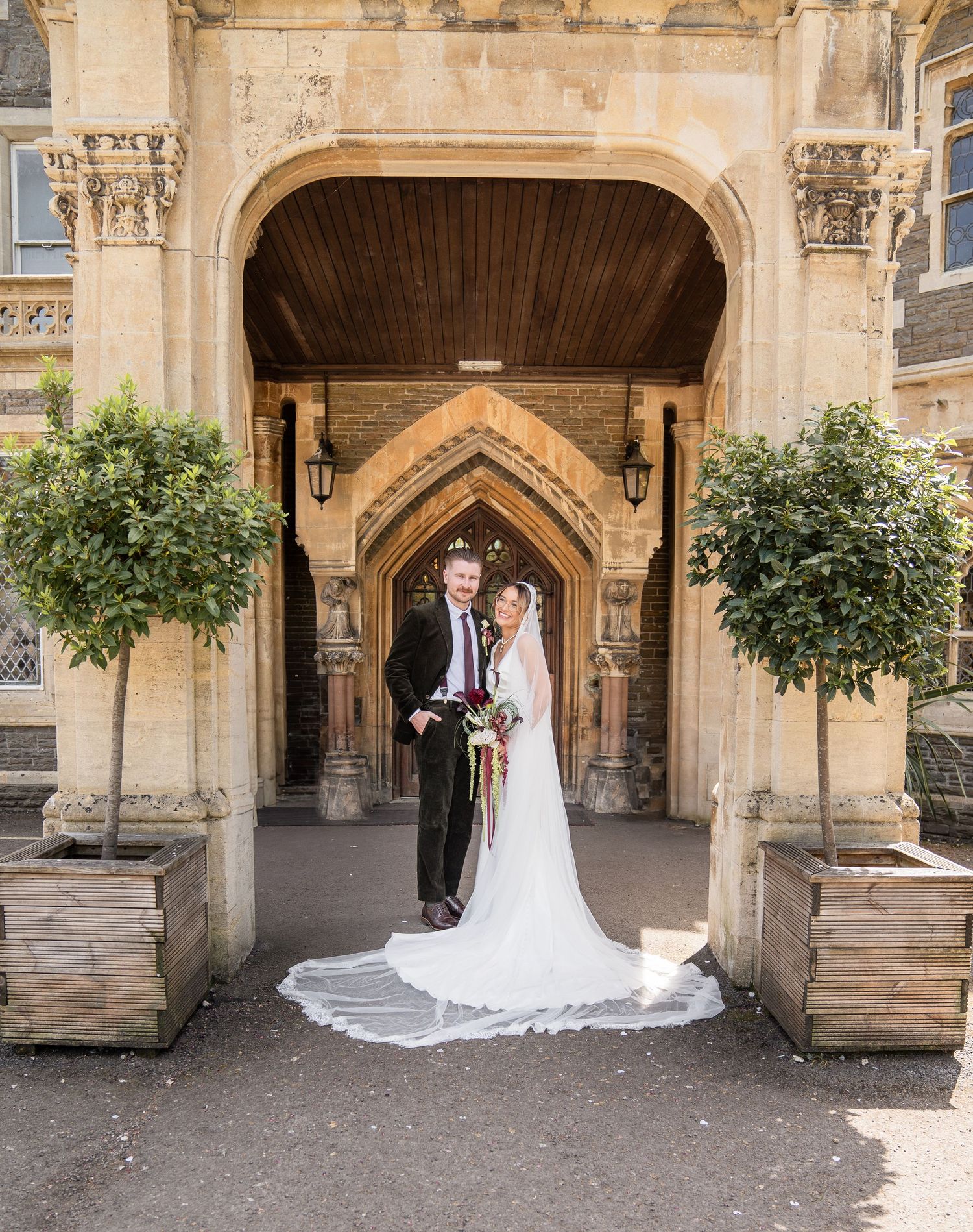 Bride and groom posing under a grand stone archway with ornate Gothic details and potted trees at a historic wedding venue.