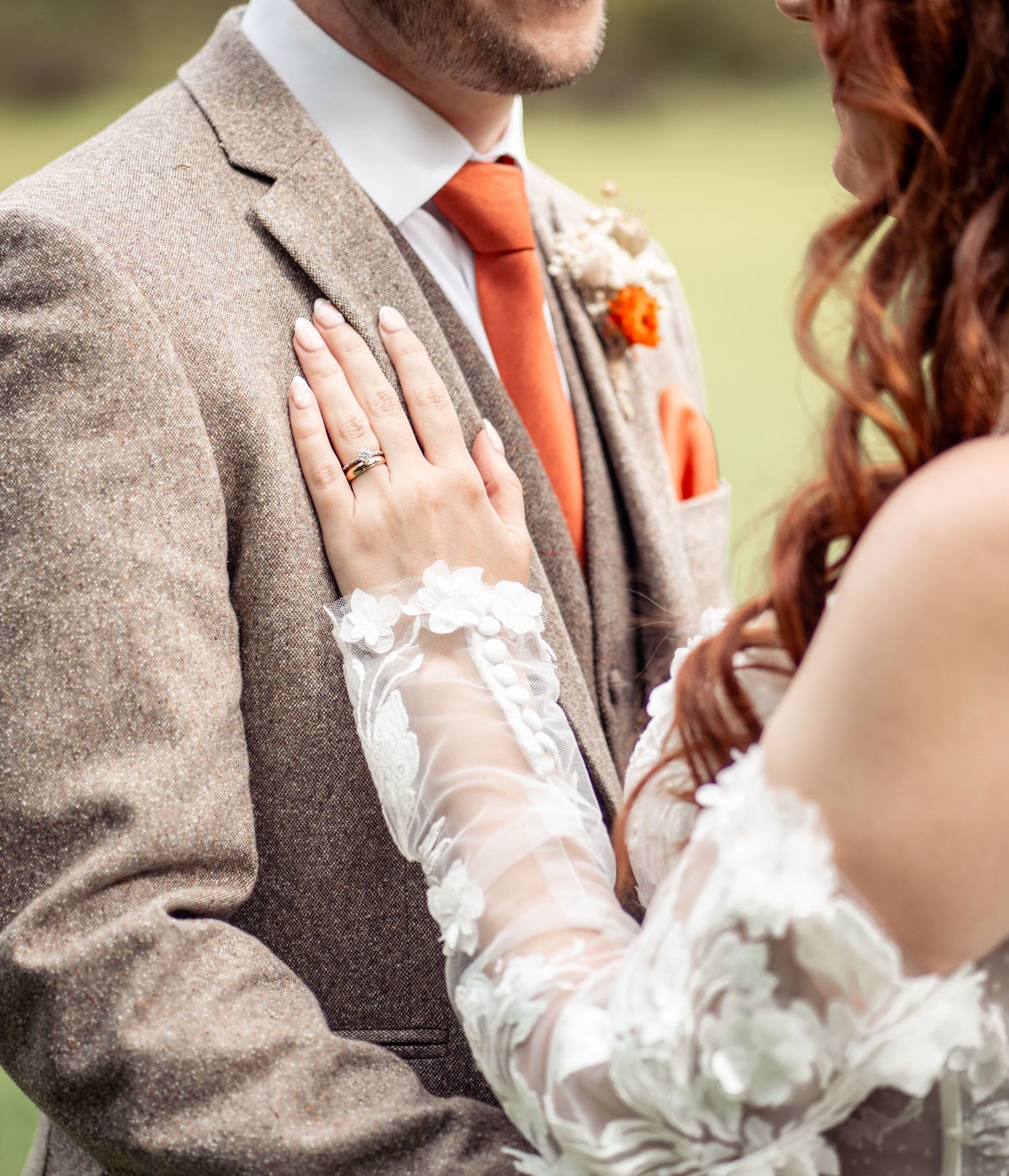 Close-up detail of a couple in wedding attire with lace sleeve and brown tweed suit with orange accents.