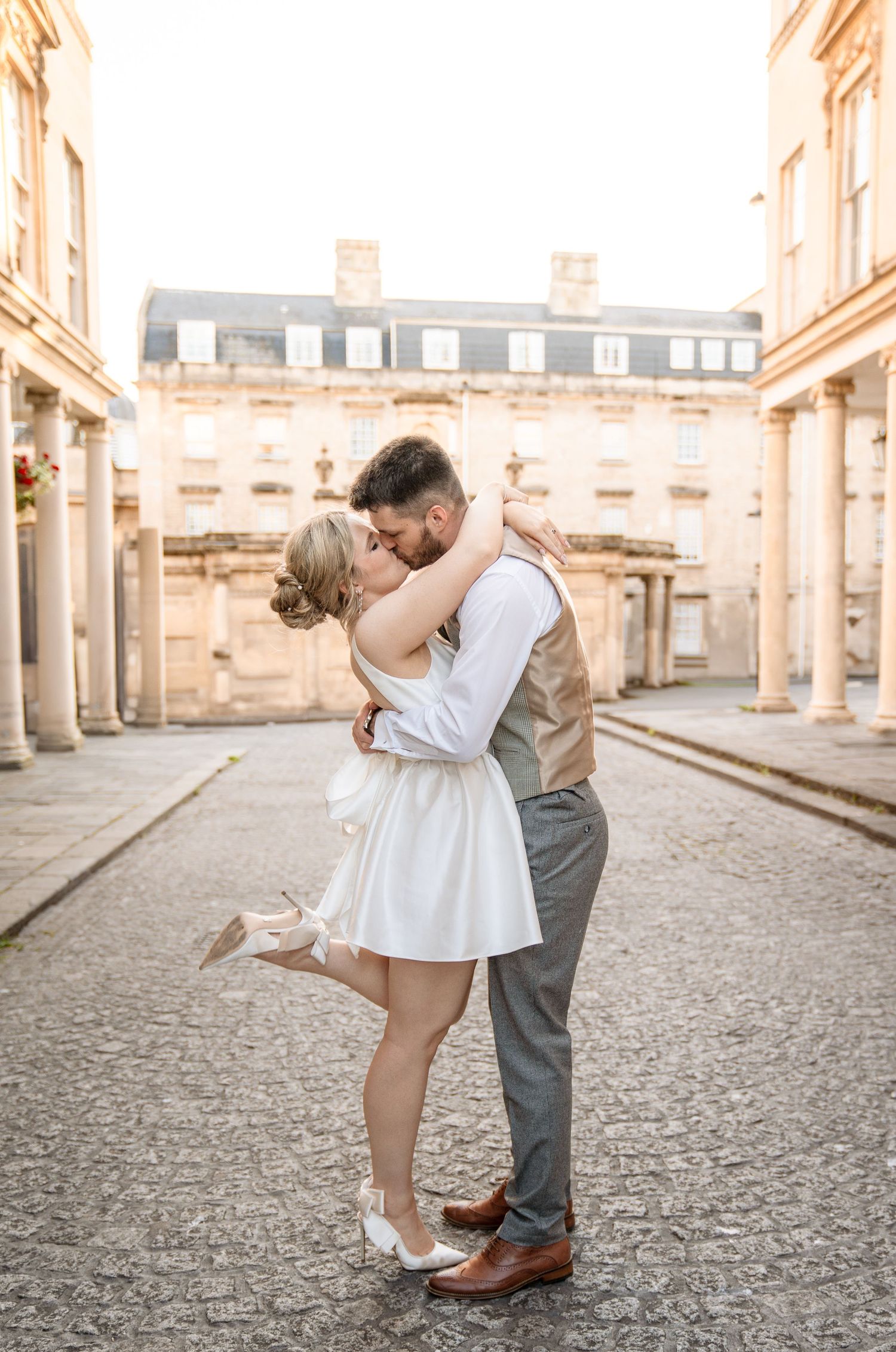 A couple shares a romantic kiss on a cobblestone street, the bride in a white mini dress lifting her foot, groom in vest and trousers.