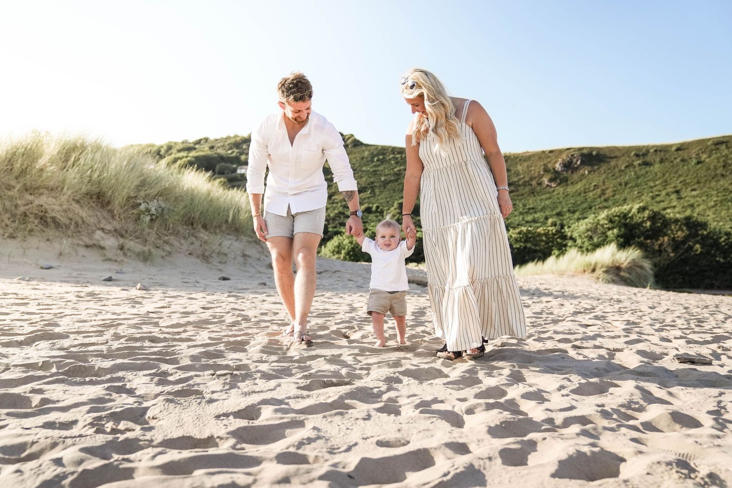 A couple in white outfits walks together on a sandy beach path during sunset.