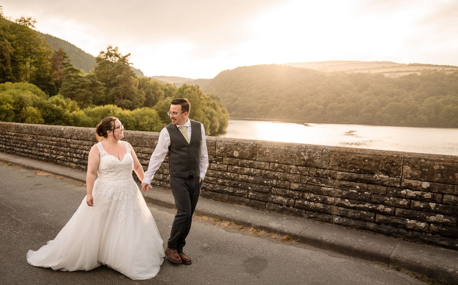 Bride and groom hold hands walking on a stone bridge with a scenic lake and lush green hills at golden hour.
