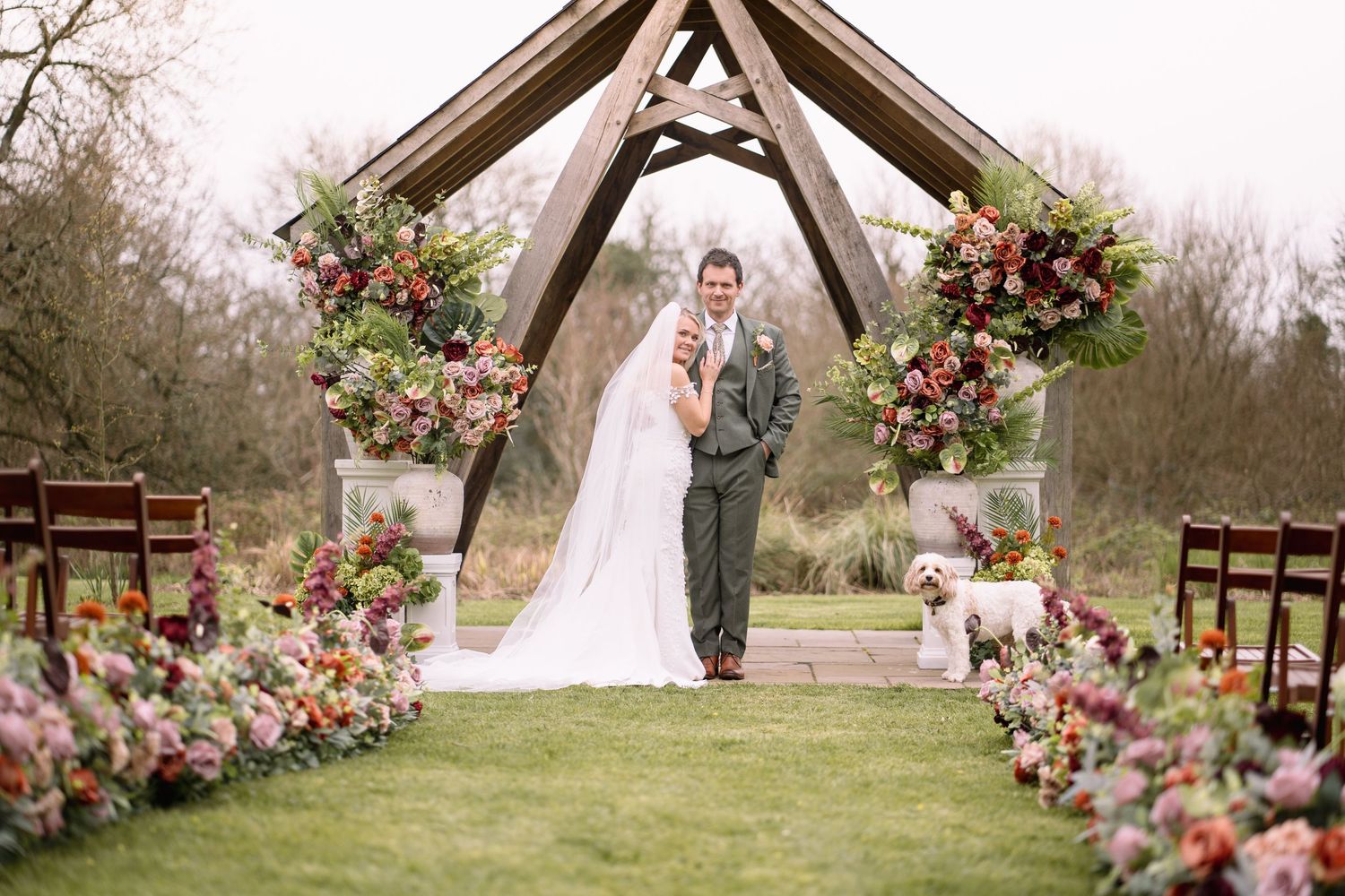 Wedding ceremony setup at outdoor venue with wooden arch decorated in colorful autumn florals and rustic wooden chairs.
