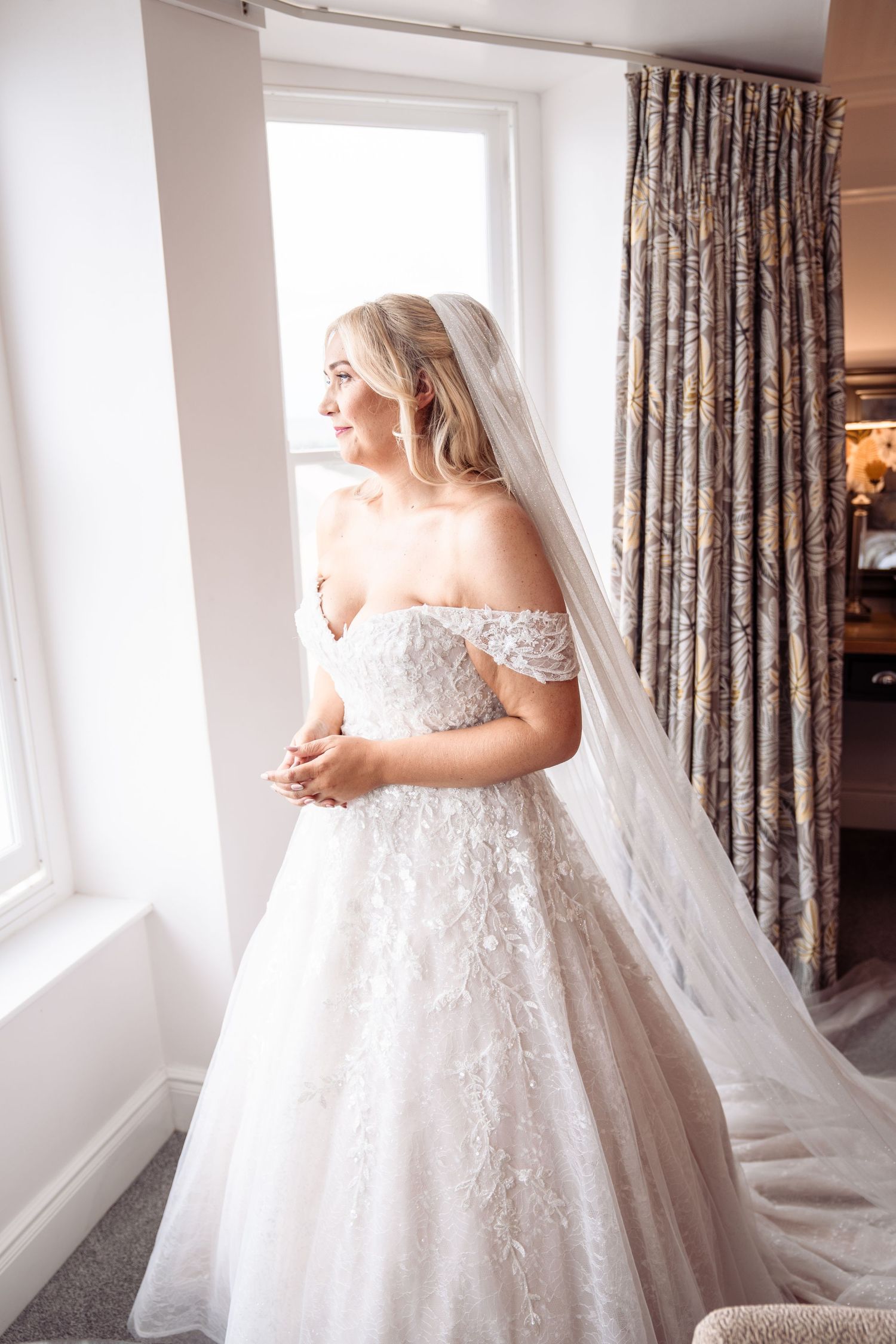 A bride in an elegant off-shoulder lace ball gown stands by a window with flowing veil in a series of photos.