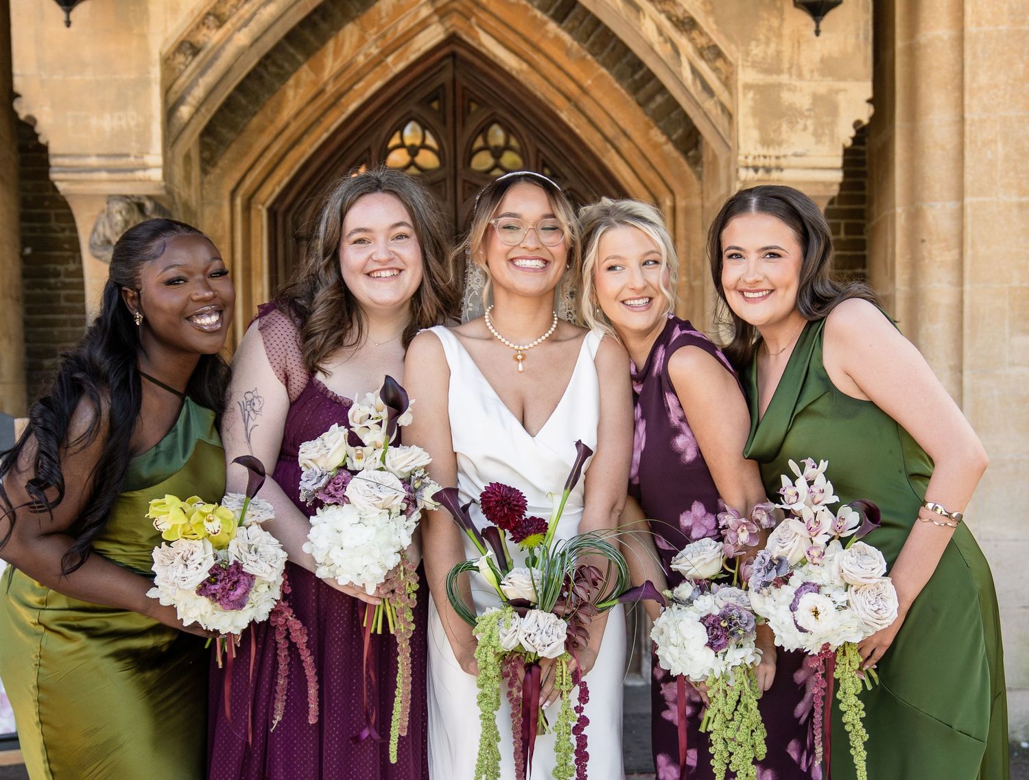 Bride smiling with four bridesmaids in green and purple dresses holding bouquets outside a stone church.