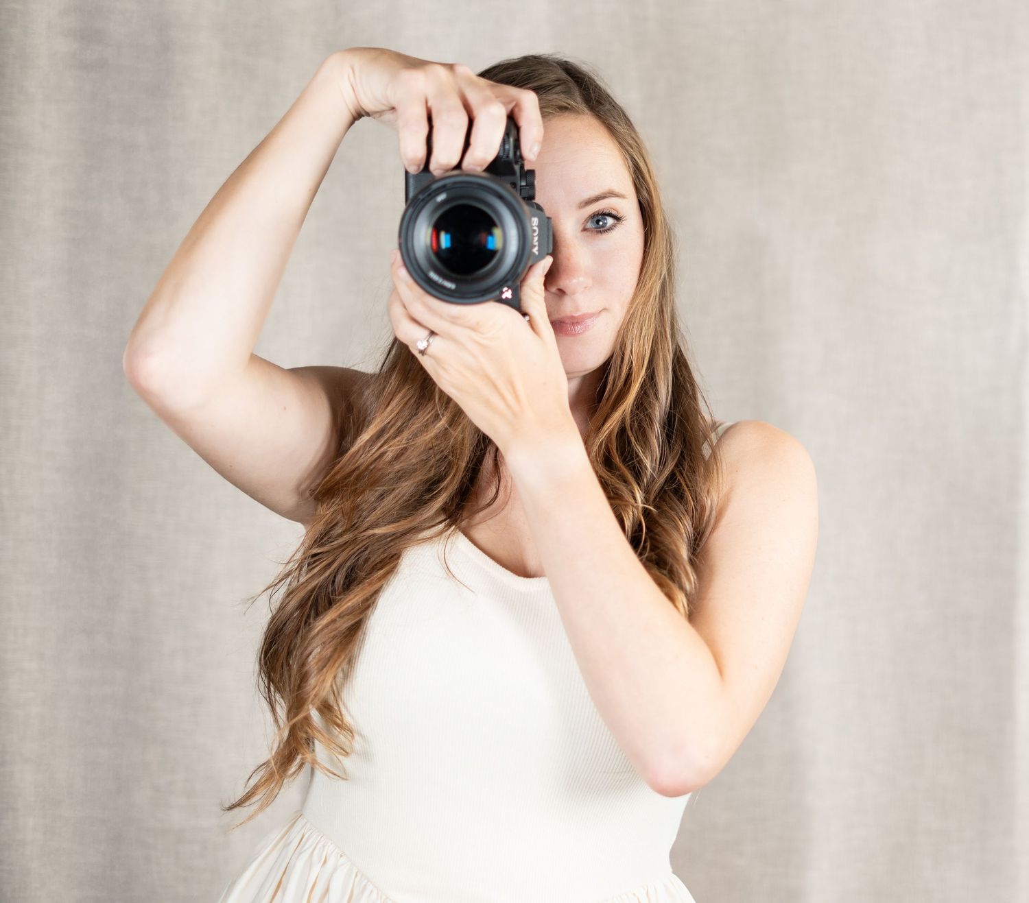 Photographer in white dress holding professional camera taking photo against light gray backdrop.