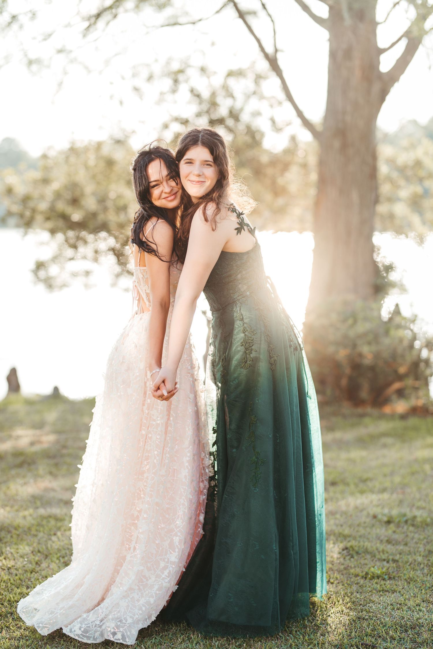 Two people in formal dresses pose together on grassy hill during golden hour.