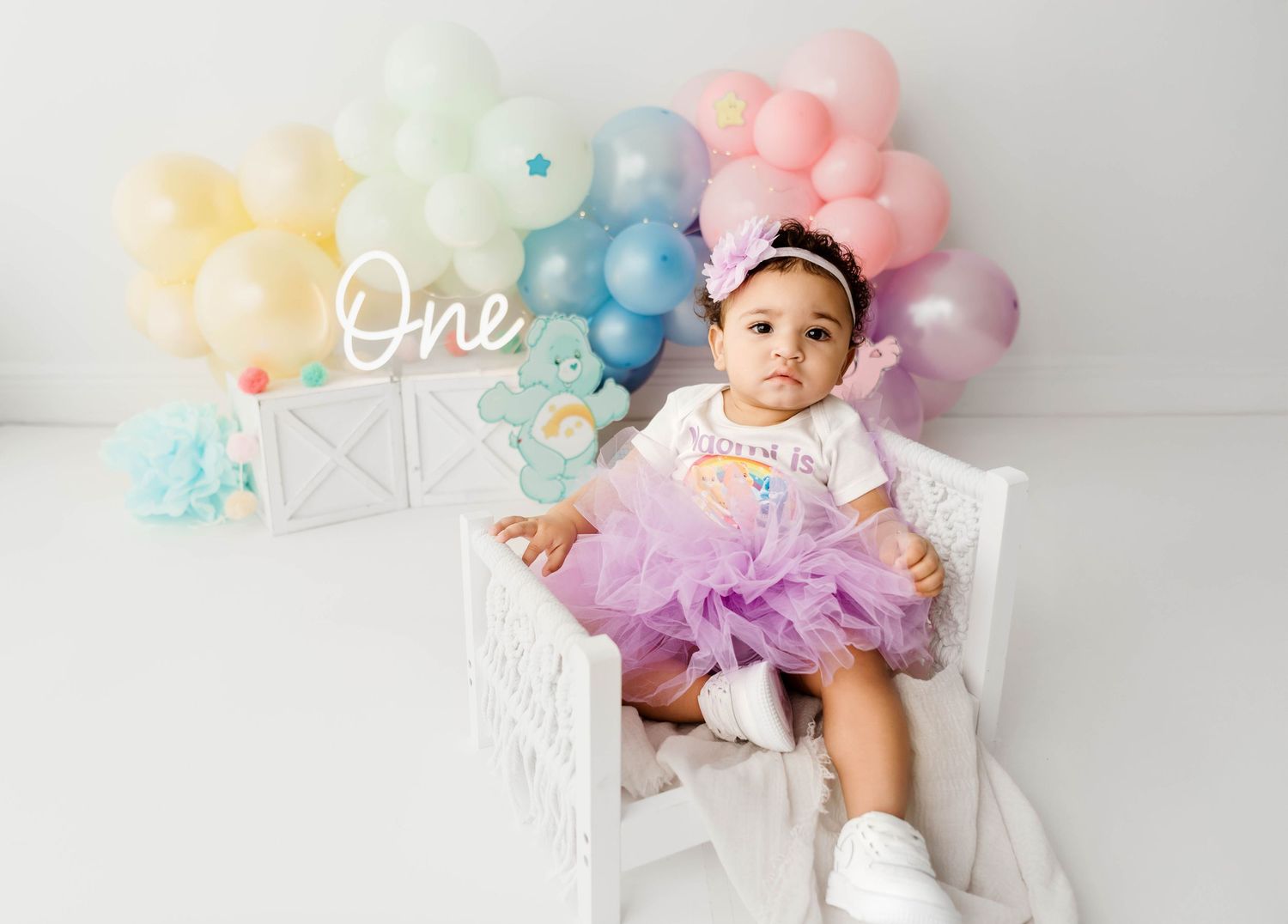 Birthday setup with white bench and pastel balloon garland against white backdrop.