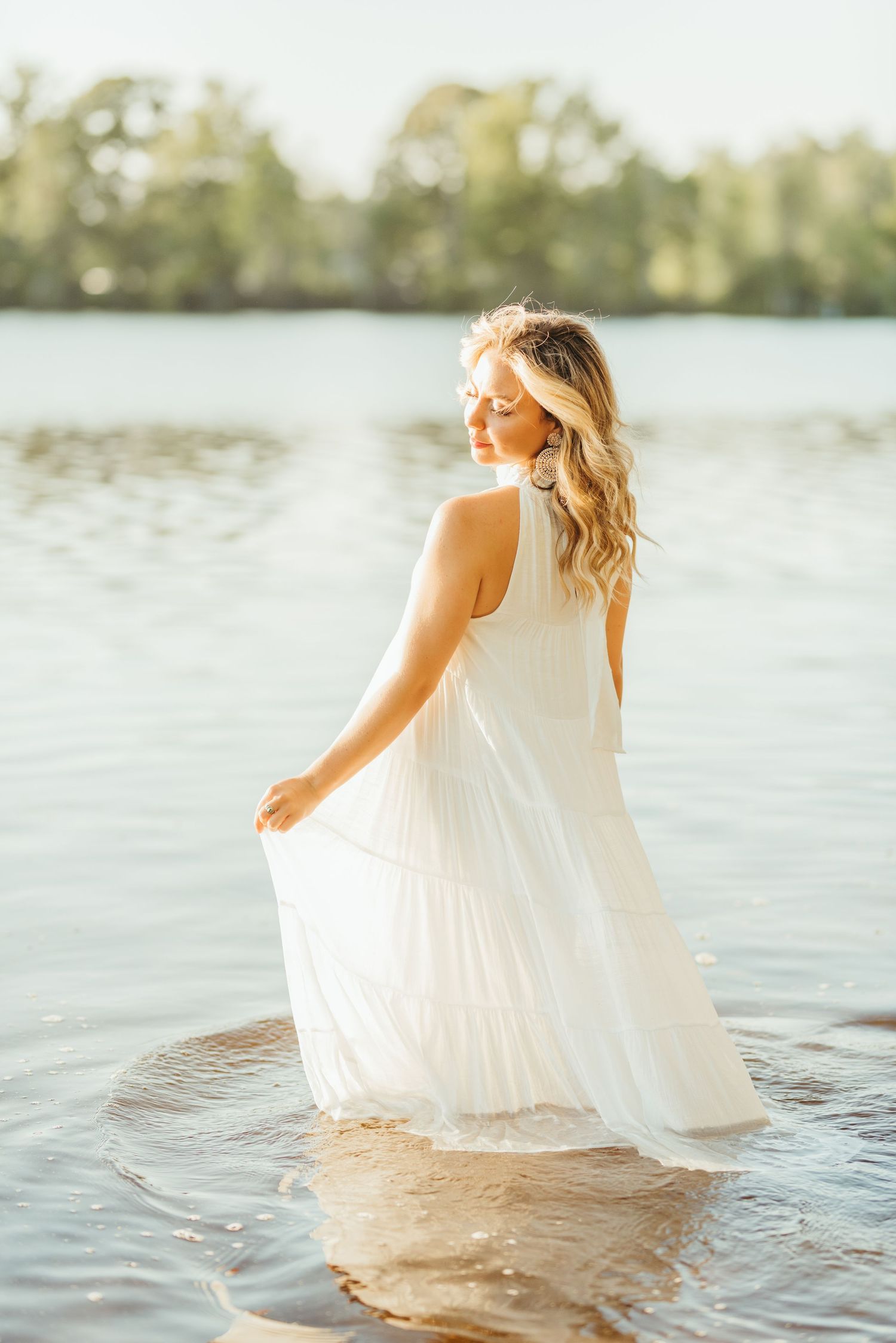 Figure in flowing white dress sitting in calm lake water during sunset.