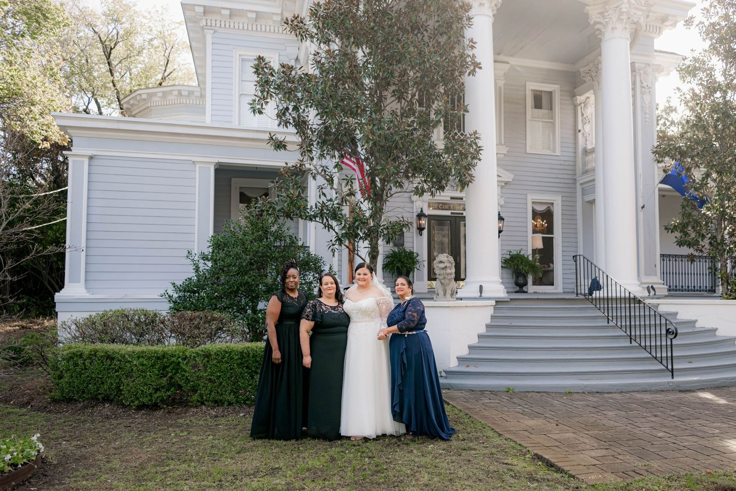 Bride in white gown with bridesmaids in navy dresses posing before a grand Southern mansion with American flag.